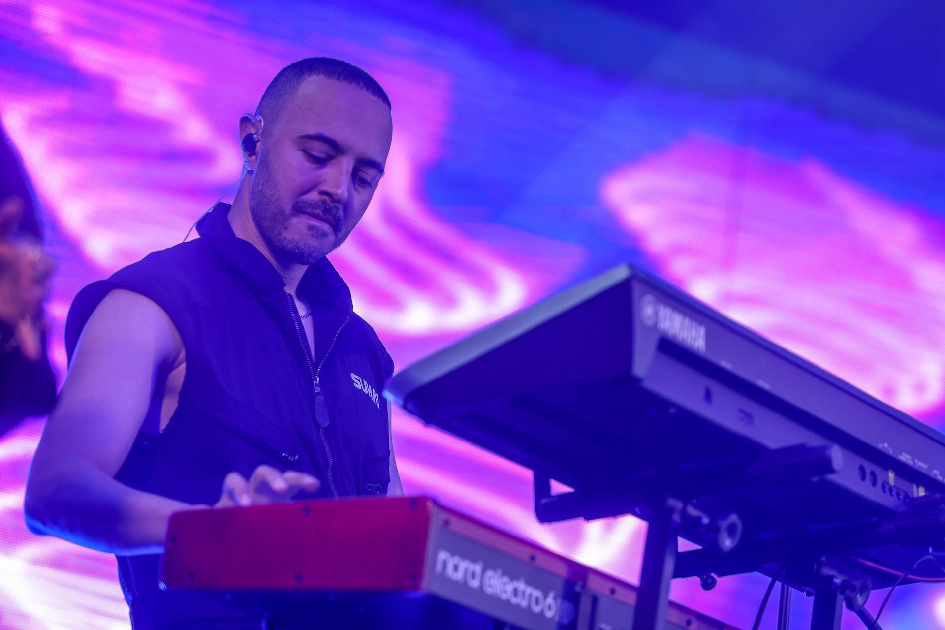 Um artista tocando um teclado Nord vermelho e um sintetizador Yamaha em um palco com um fundo abstrato ondulado na cor roxa.