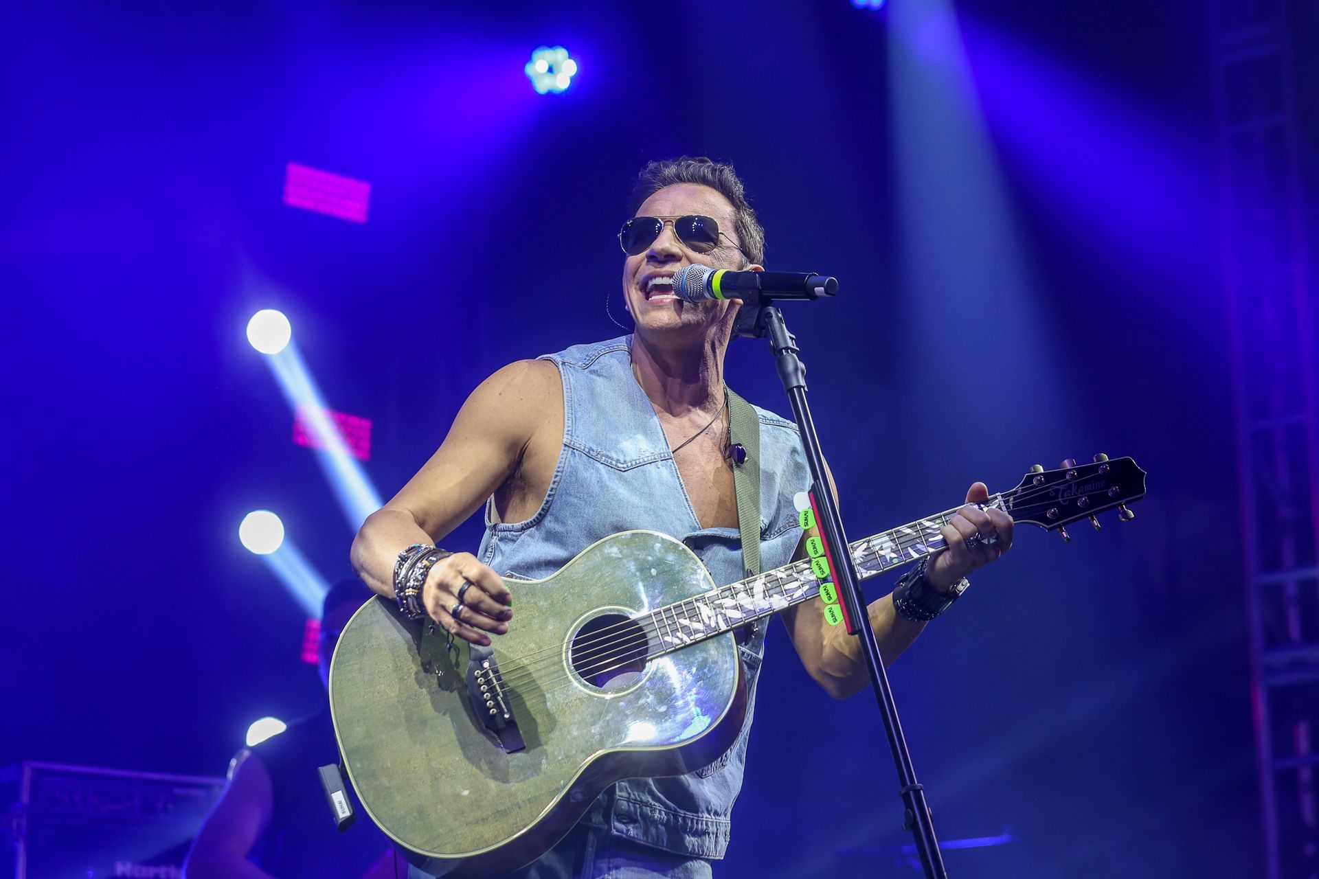 Um músico de óculos escuros e colete jeans cantando e tocando violão em um palco com iluminação azul brilhante.