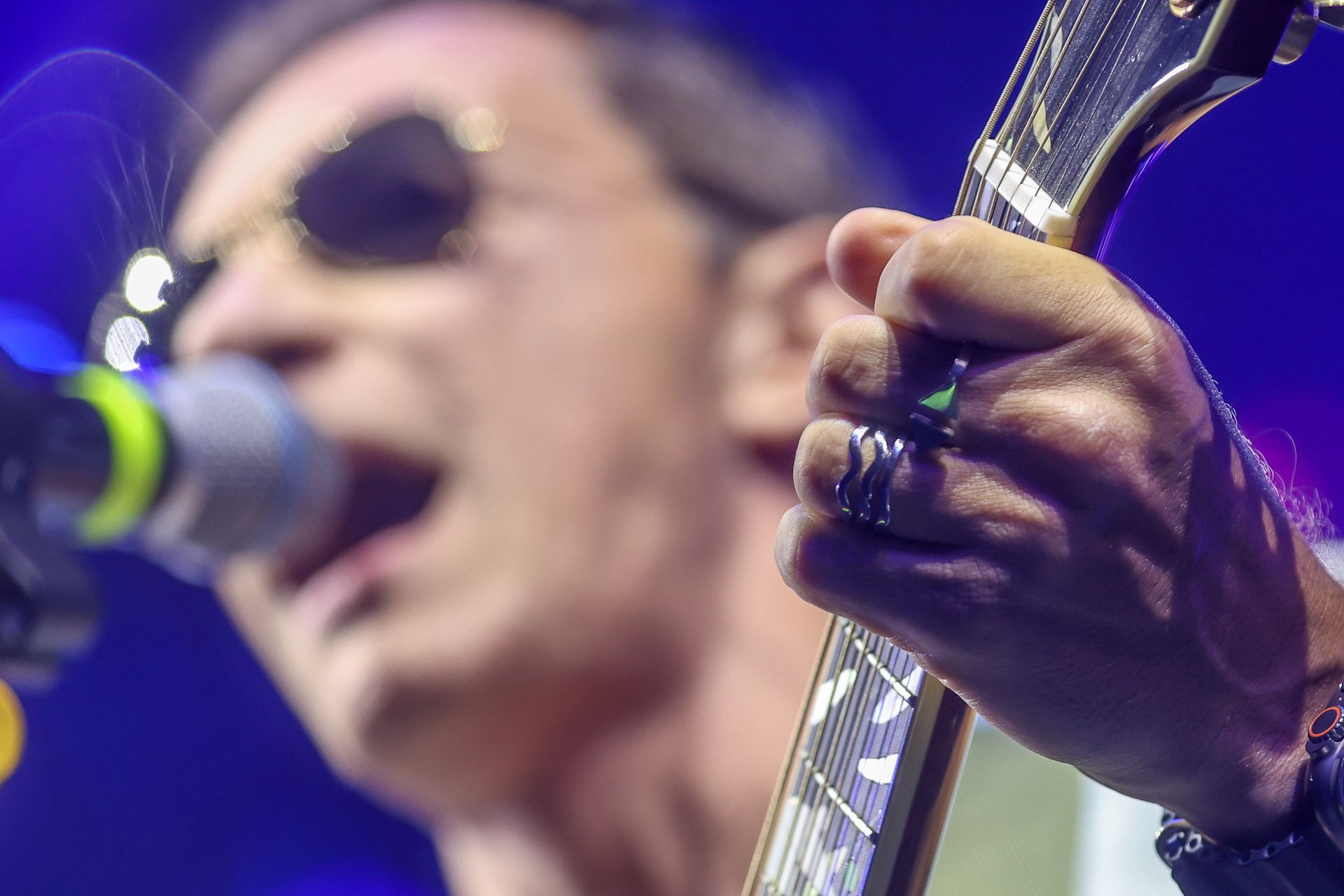 Primeiro plano de um músico usando óculos escuros tocando guitarra elétrica enquanto canta em um microfone no palco.
