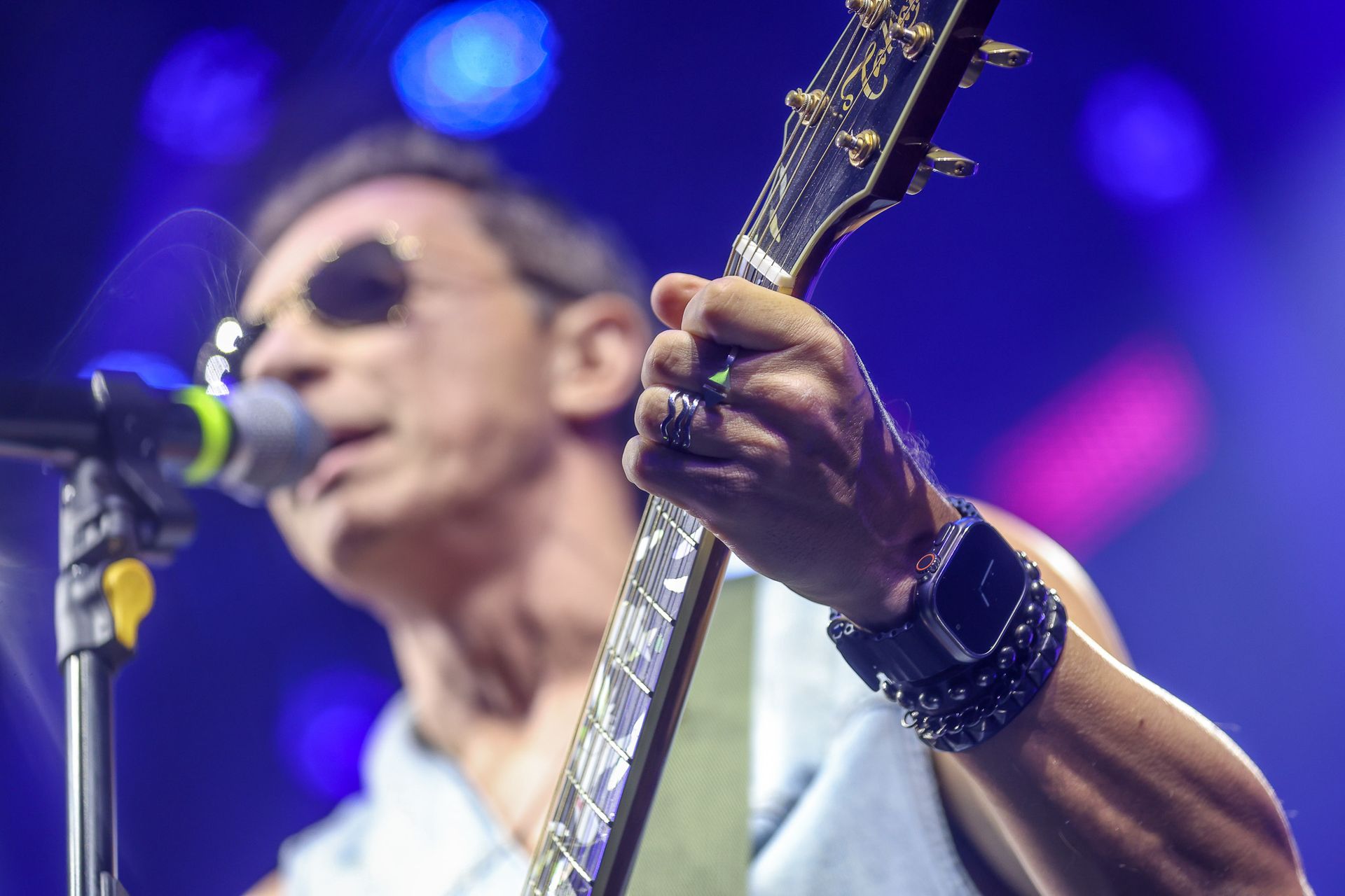 Um músico usando óculos escuros toca uma guitarra elétrica com escala decorada enquanto se apresenta no palco.