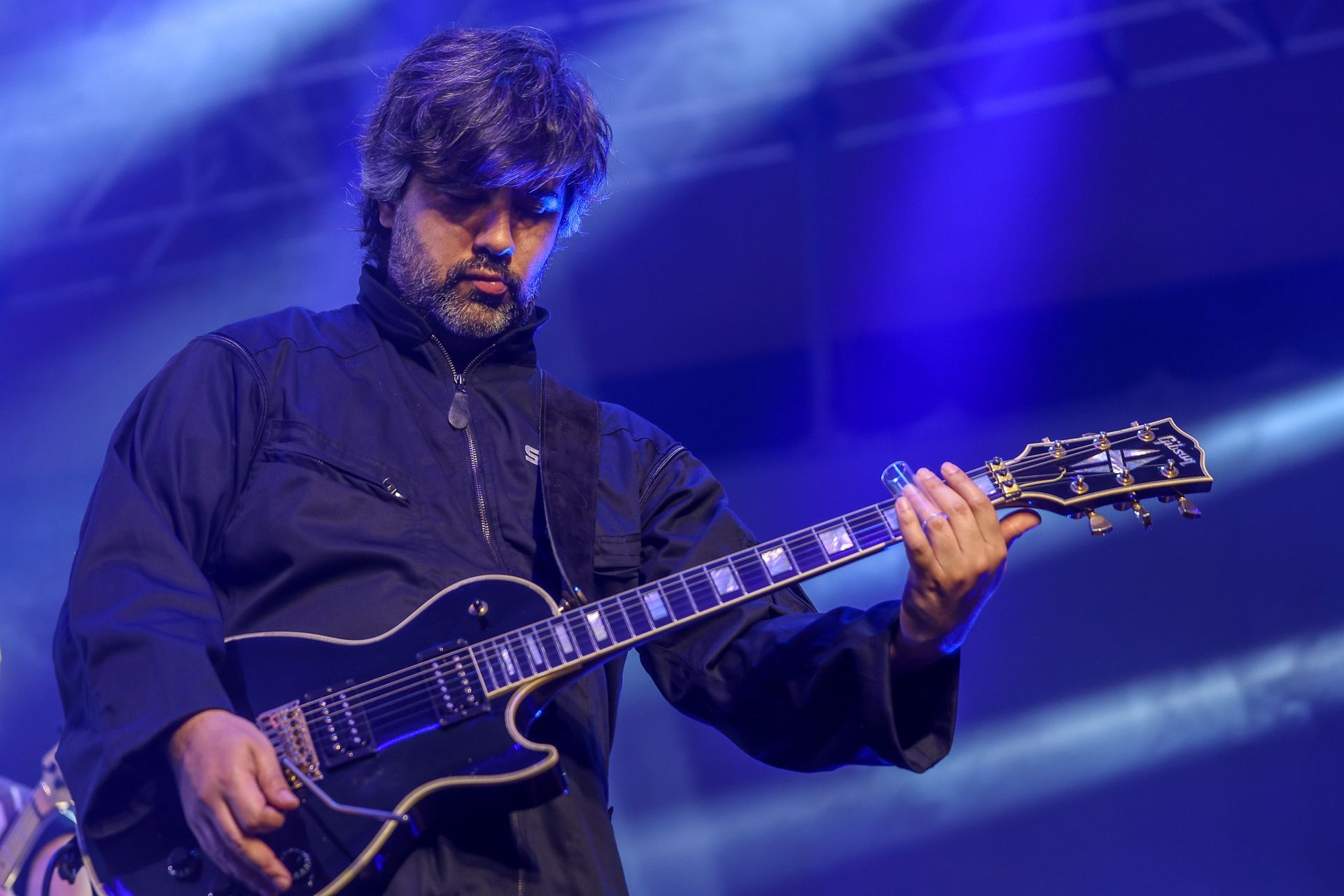 Um músico de jaqueta preta tocando guitarra elétrica preta no palco sob luzes azuis de concerto.
