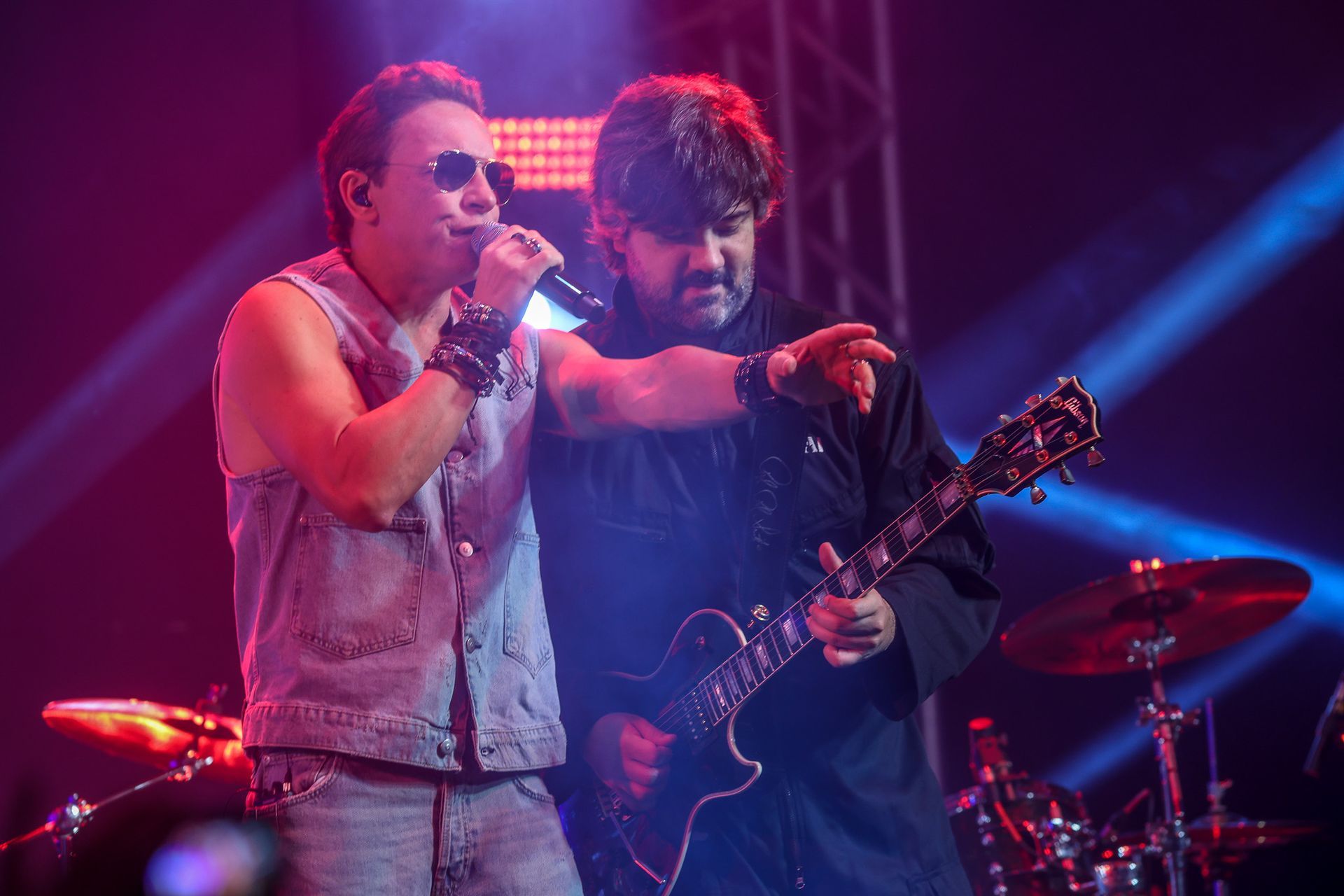 O vocalista, de óculos escuros e colete jeans, enquanto um guitarrista toca próximo a ela em um palco iluminado por luz azul.