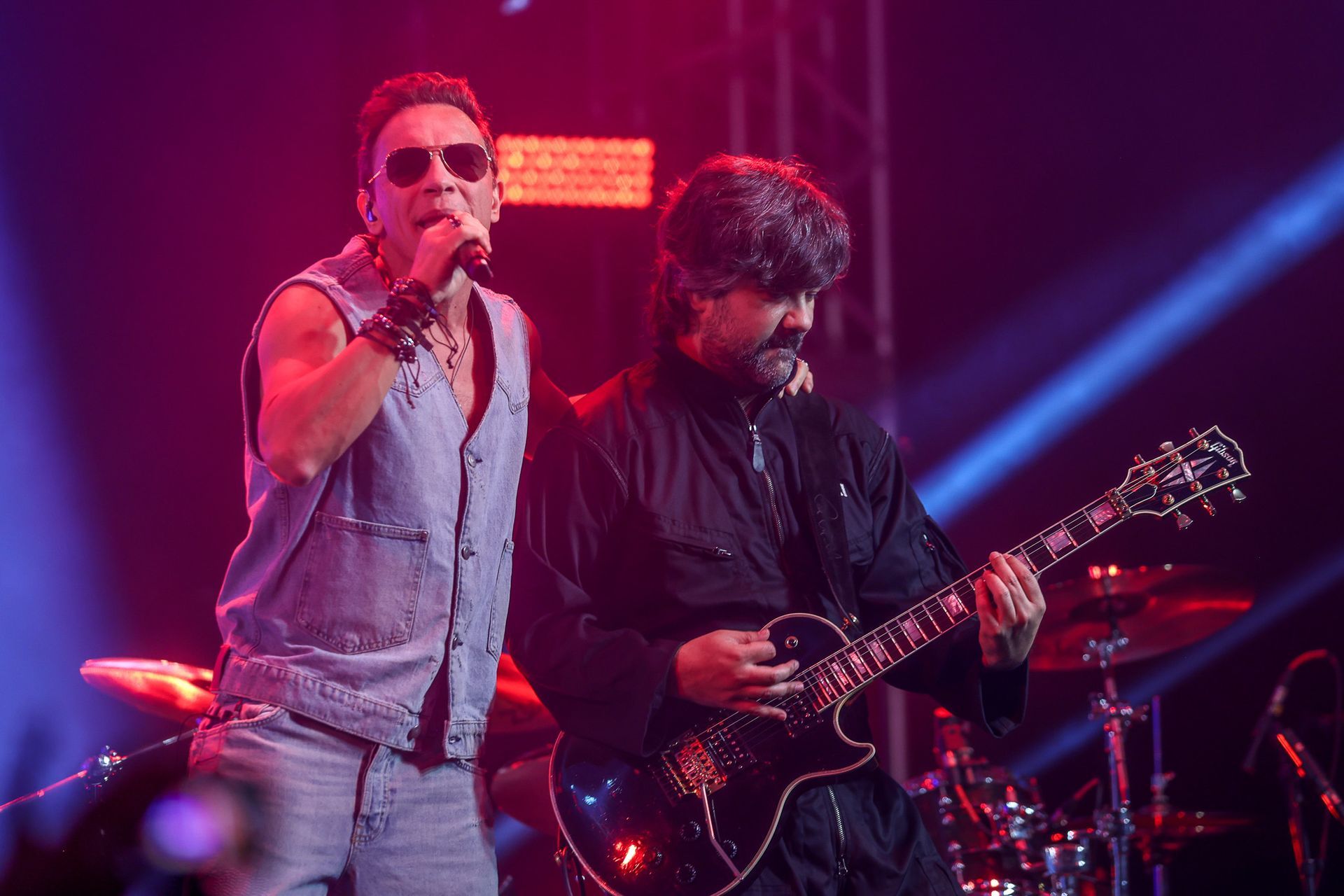 Um cantor de colete jeans e óculos escuros se apresenta com um guitarrista no palco.