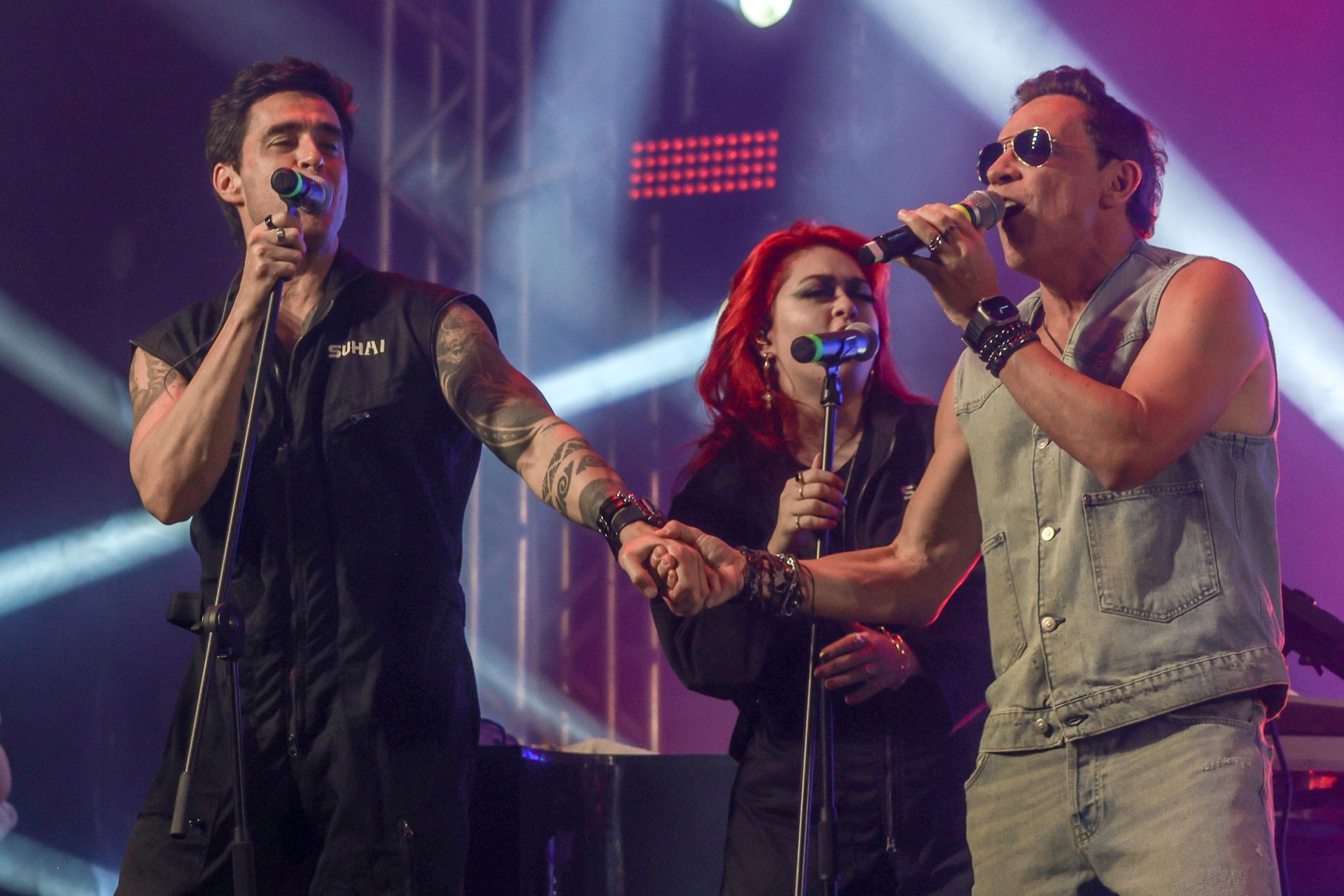 Três pessoas cantando em um palco de shows, com dois homens de mãos dadas enquanto se apresentam sob luzes fortes.