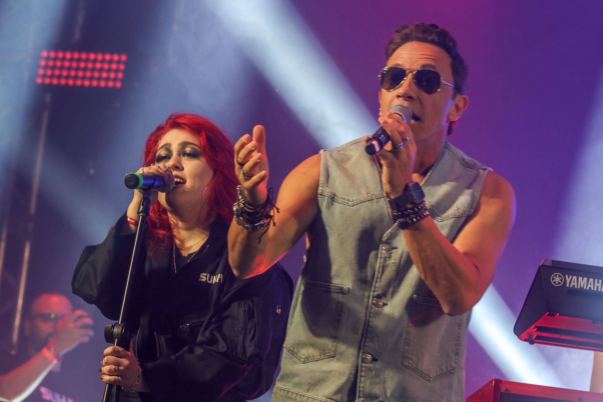 Dois artistas no palco cantando em microfones, uma com cabelo ruivo e jaqueta preta, o outrousando óculos escuros.