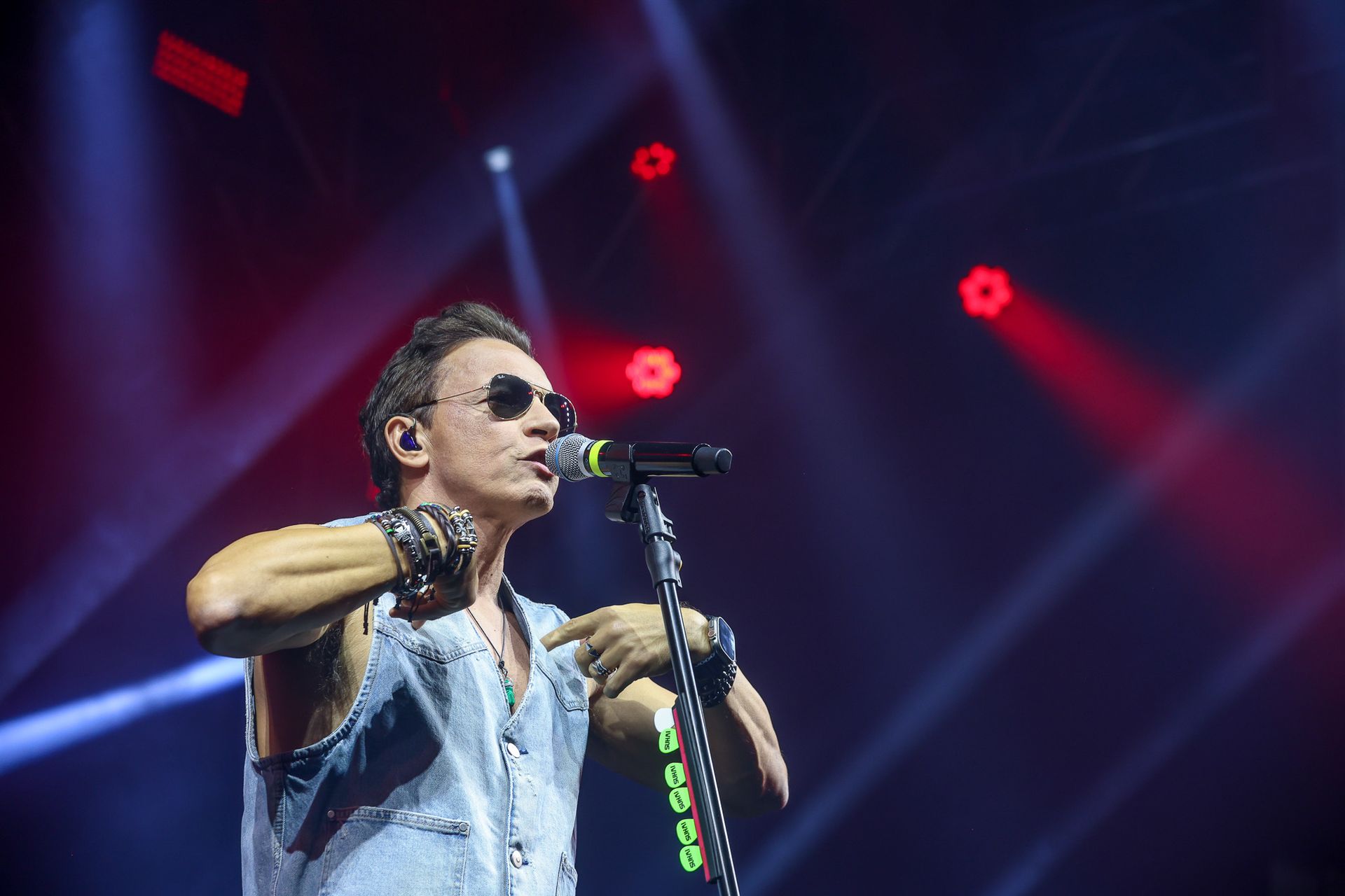 Um cantor vestindo colete jeans e óculos escuros se apresenta no palco com luzes vermelhas brilhando ao fundo.
