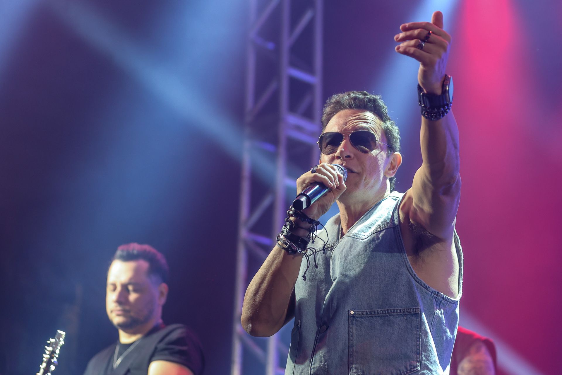 Um cantor de óculos escuros no palco falando ao microfone, enquanto um músico toca violão atrás dele.