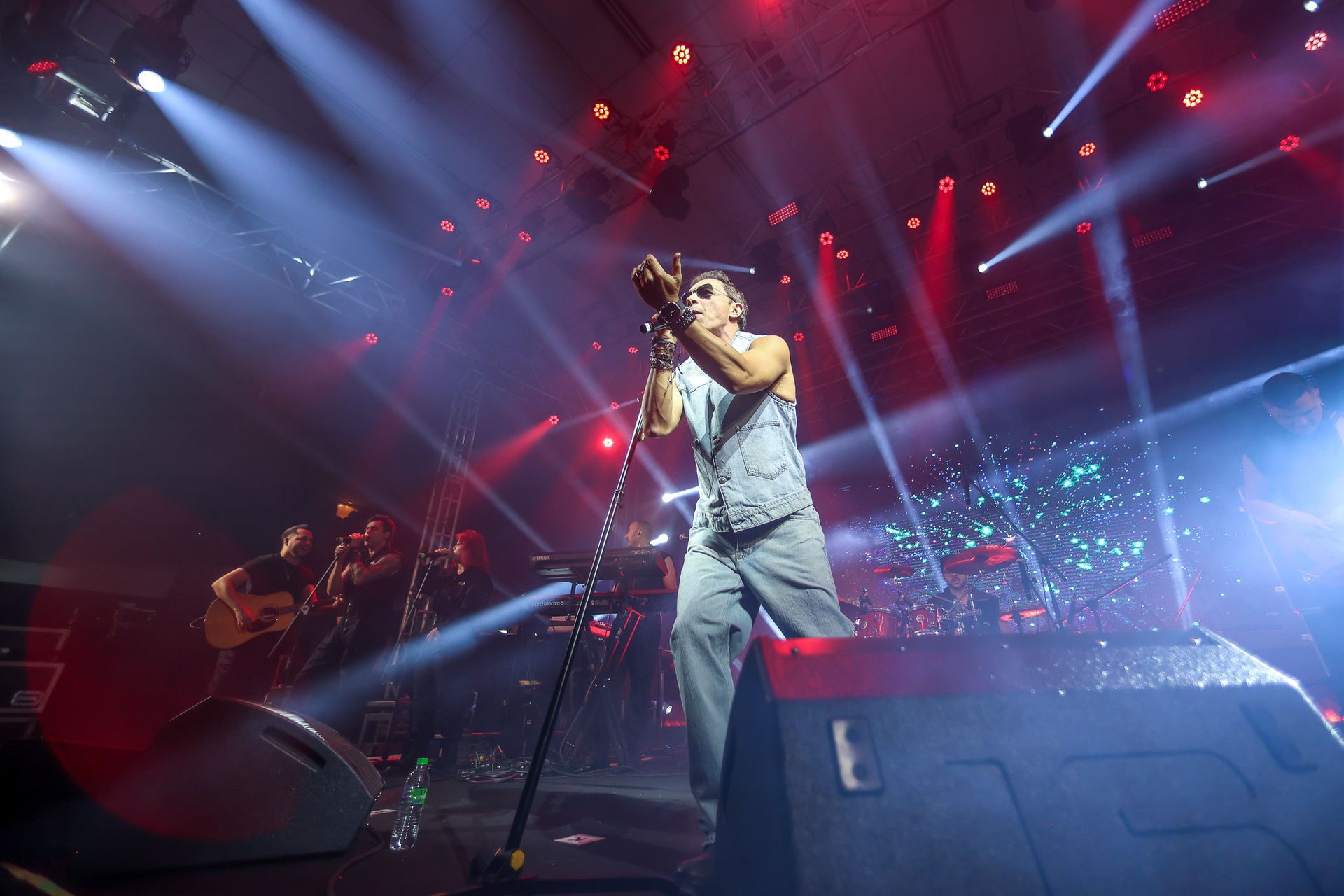Uma vocalista se apresenta no palco, falando ao microfone sob holofotes de um show, com os membros da banda por perto.