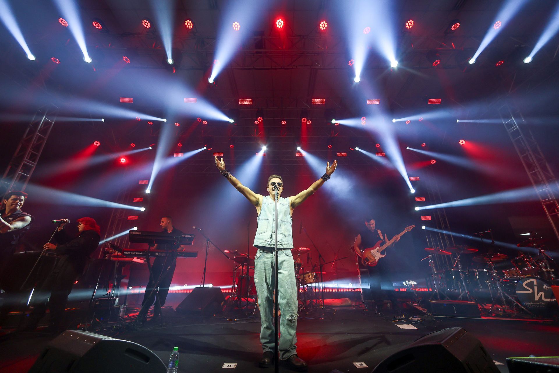 Um cantor está de pé no centro do palco de um concerto, com os braços erguidos sob luzes dramáticas vermelhas e brancas.