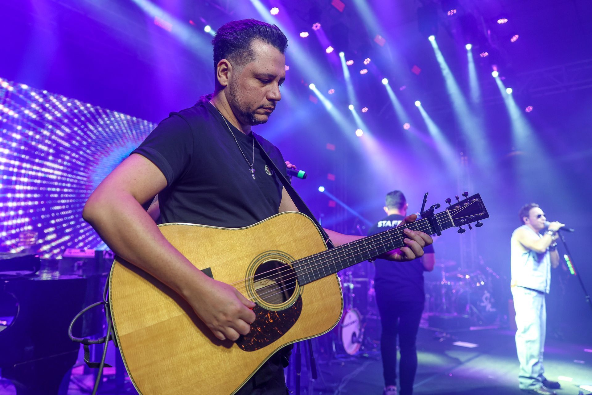 Um músico toca violão em um palco banhado por luzes azuis e roxas, com os membros da banda tocando nas proximidades.