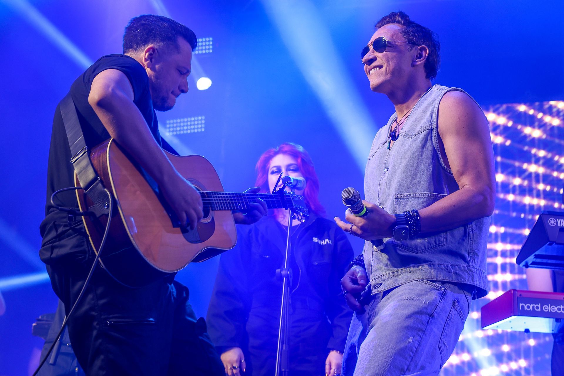 Dois músicos se apresentam em um palco sob luzes azuis; um toca violão enquanto o outro canta ao microfone.