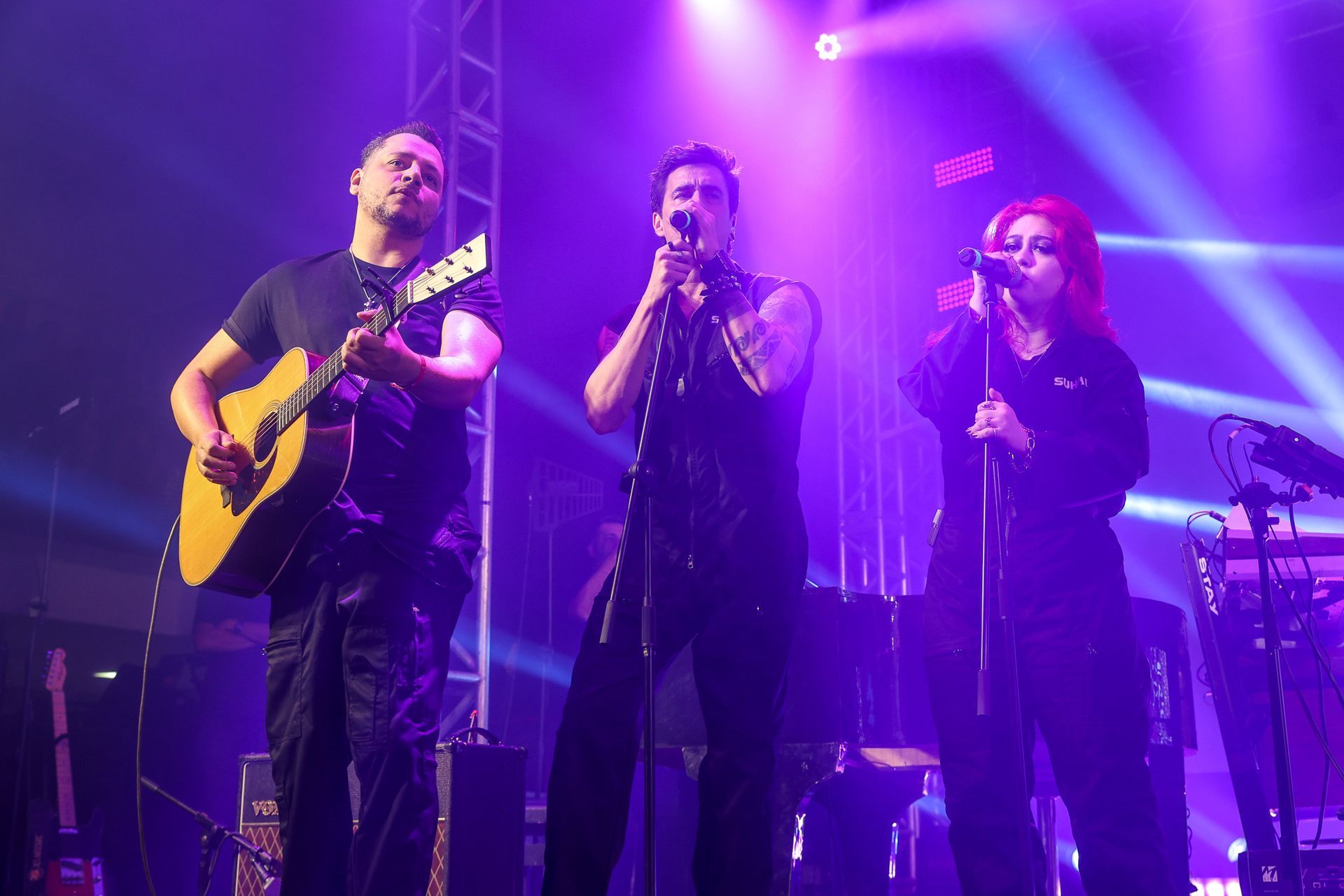 Três músicos se apresentam em um palco iluminado por holofotes roxos, um tocando violão e dois cantando em microfones.