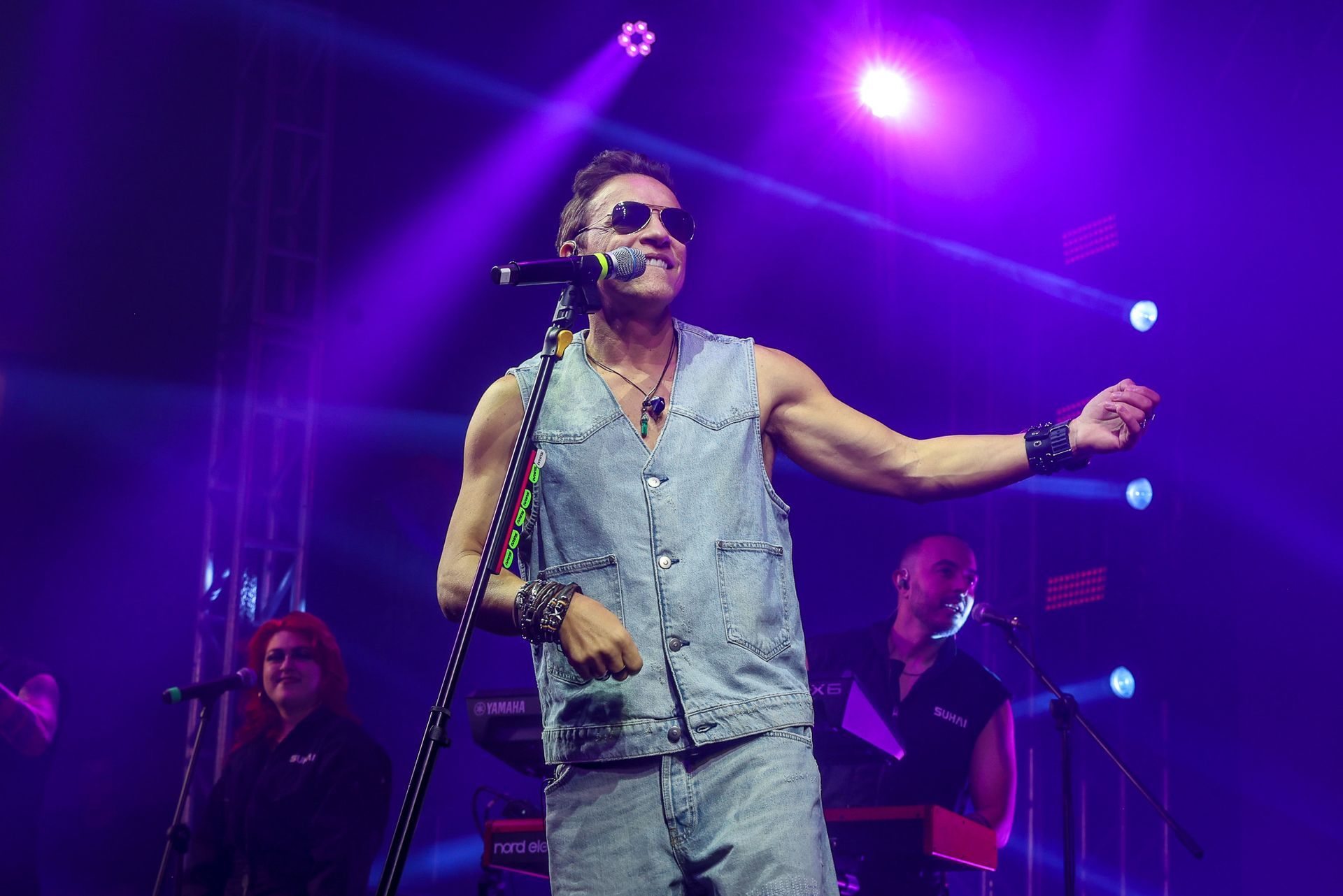 Uma pessoa vestindo um colete jeans e óculos escuros canta em um microfone em um palco iluminado por luzes roxas brilhantes.