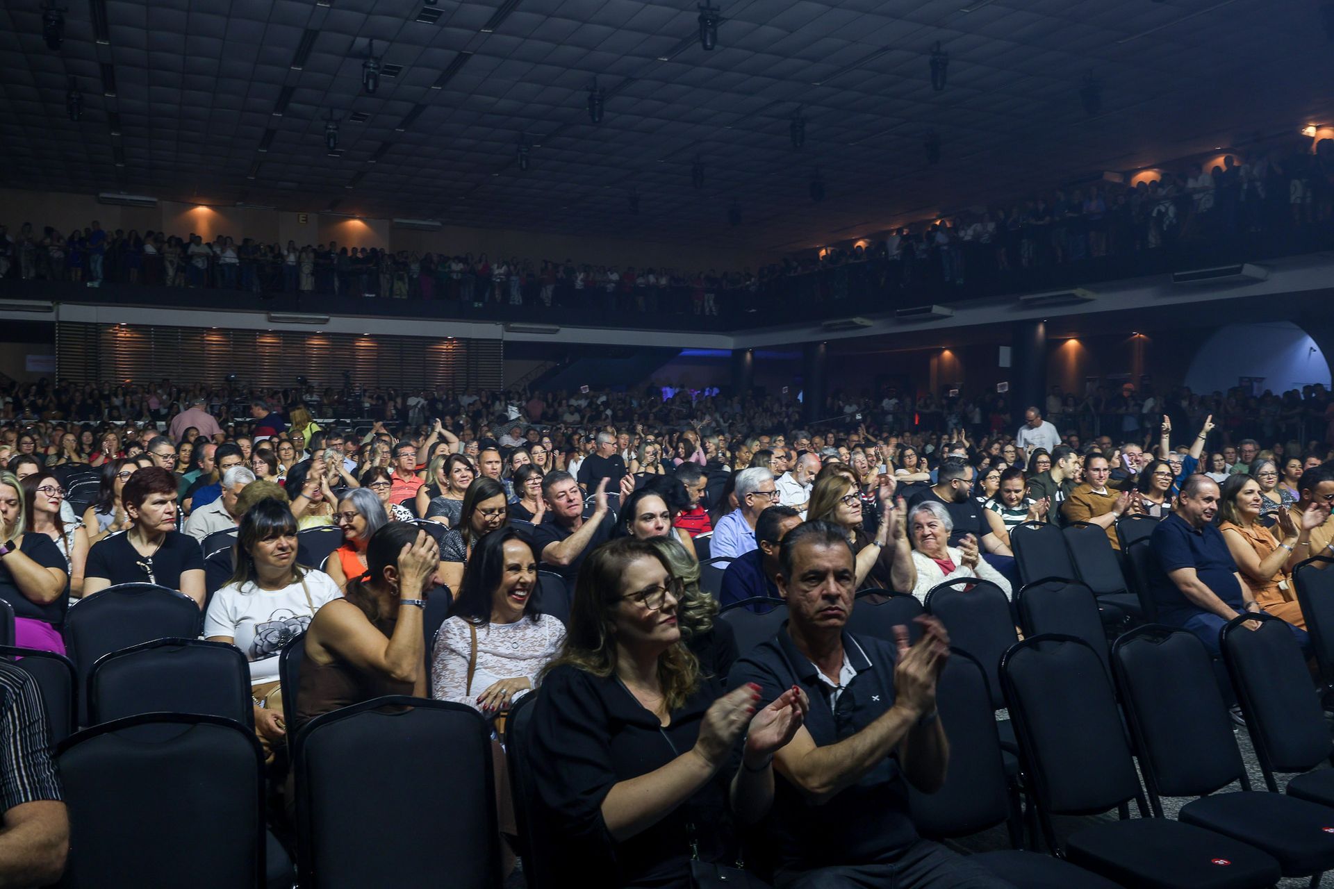 Um grande auditório escuro, por uma plateia que aplaude, sentada em fileiras de cadeiras pretas de frente para o palco.