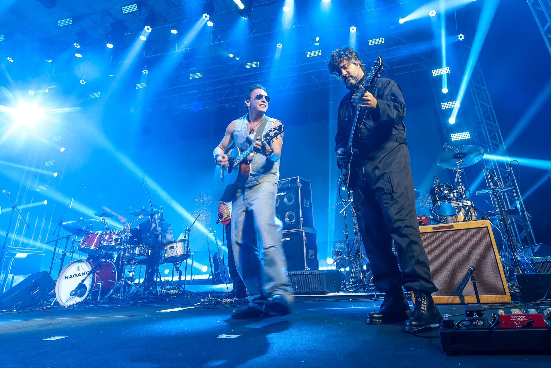 Dois músicos se apresentam em um palco de concertos banhado por uma luz, tocando guitarras em meio a equipamentos de palco.