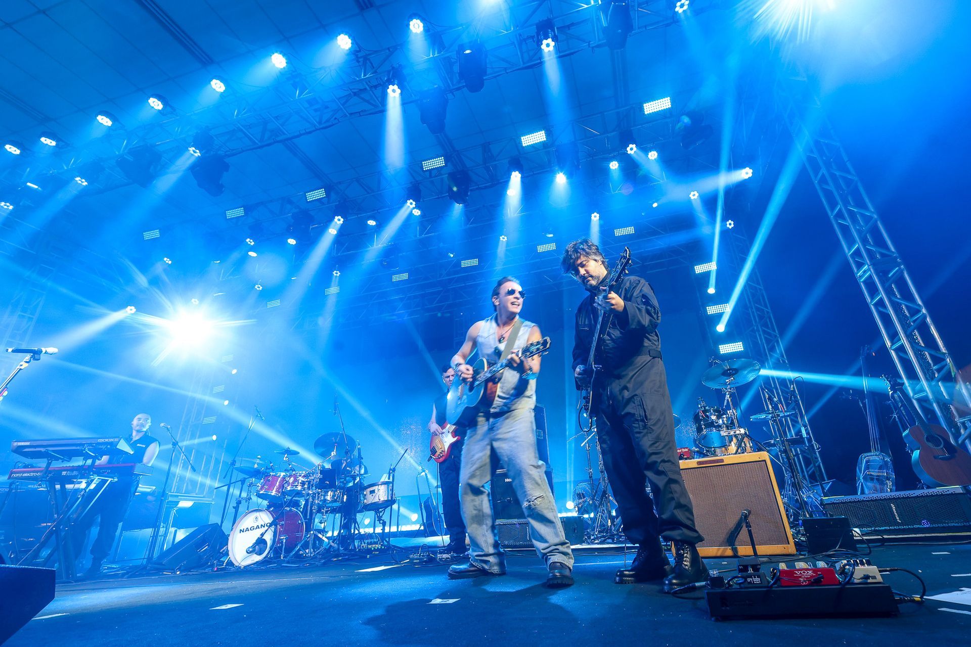 Dois músicos se apresentam no palco banhados por luzes azuis brilhantes de um show, um tocando guitarra e o outro cantando.
