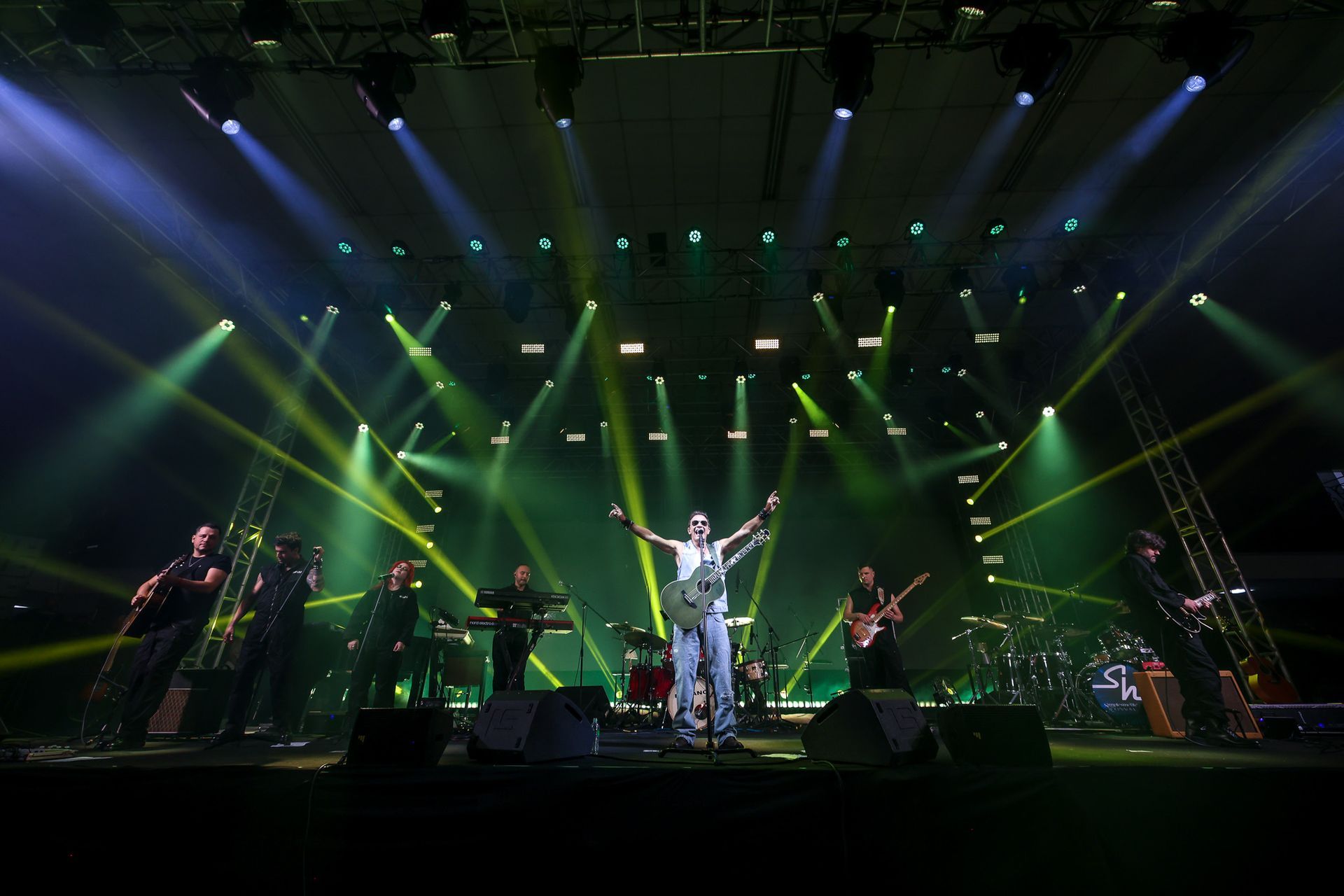 Uma banda de rock se apresenta no palco sob luzes verdes brilhantes, com o vocalista segurando uma guitarra com os braços.