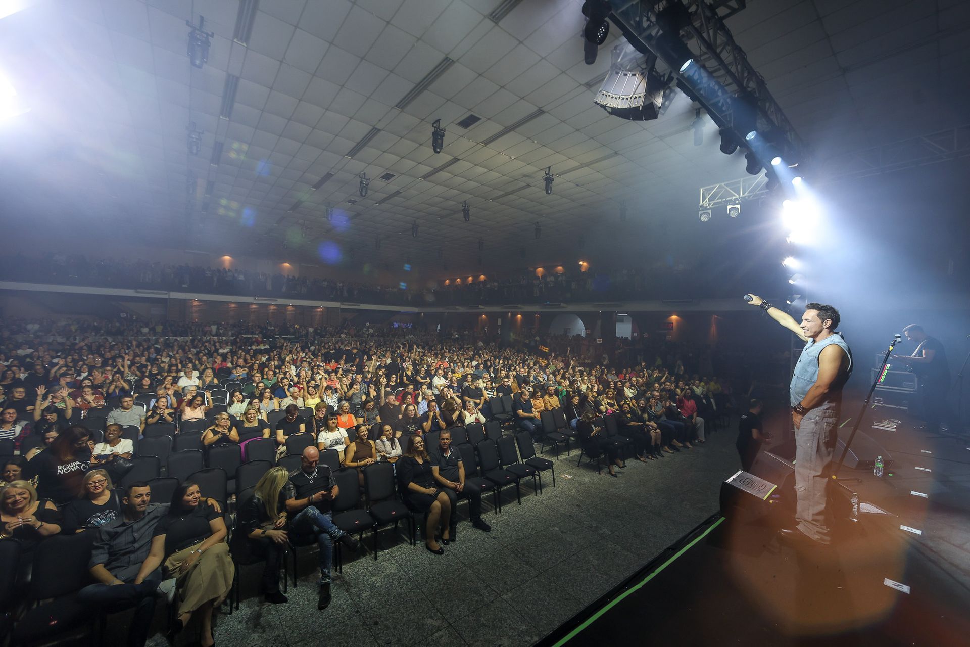 Cantor está em pé no palco gesticulando para uma grande plateia sentada em uma sala de concertos de atmosfera intimista.