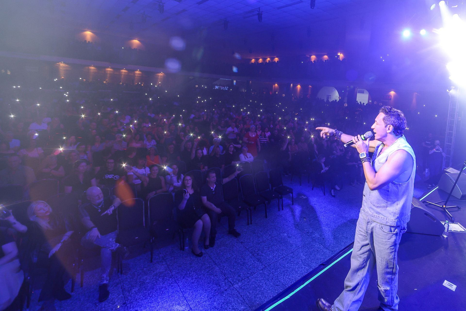 Um cantor se apresentando no palco, apontando para uma plateia iluminada pelas luzes brilhantes de celulares com pouca luz.