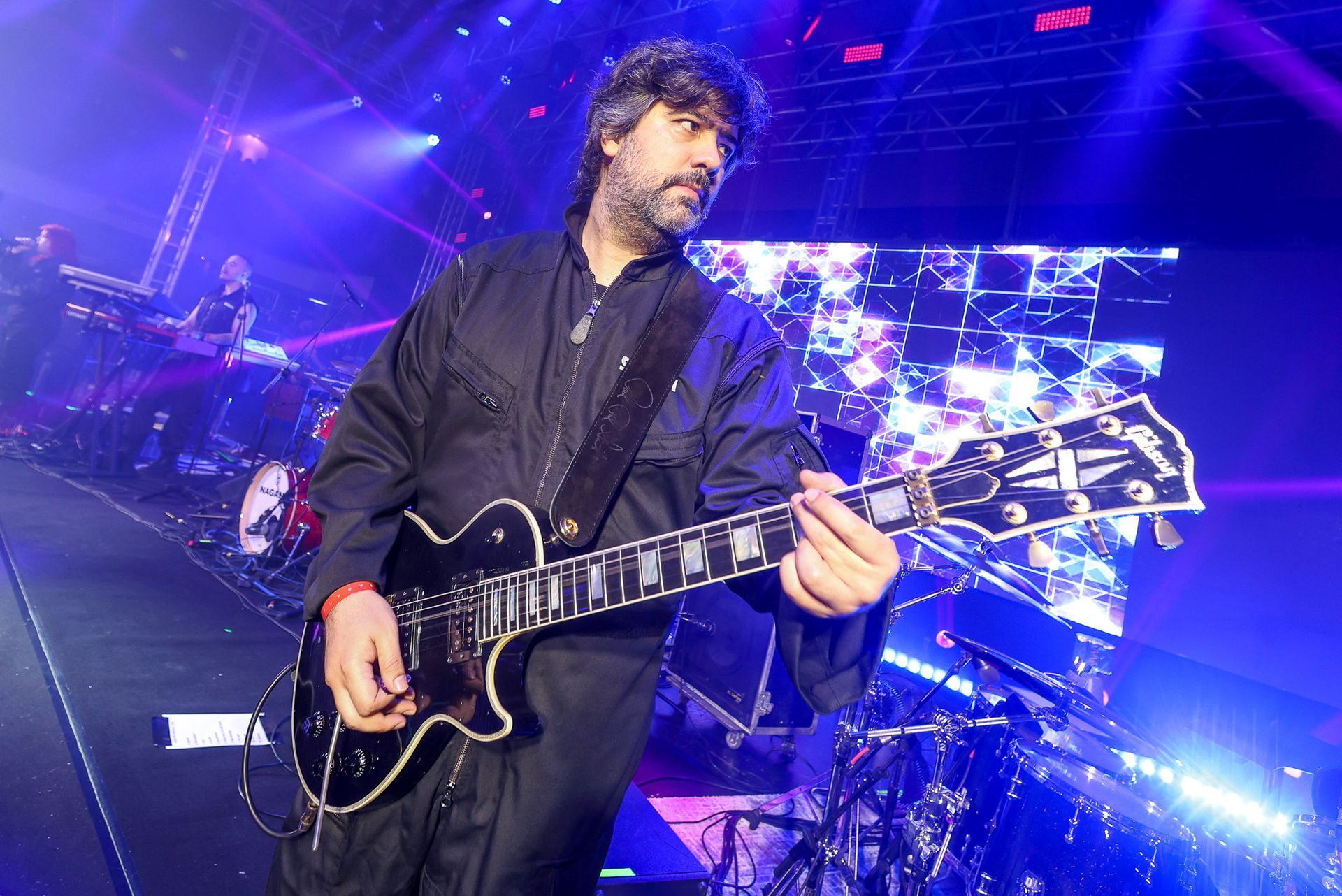 Um músico tocando uma guitarra elétrica preta em um palco iluminado com luzes azuis e uma tela quadriculada ao fundo.