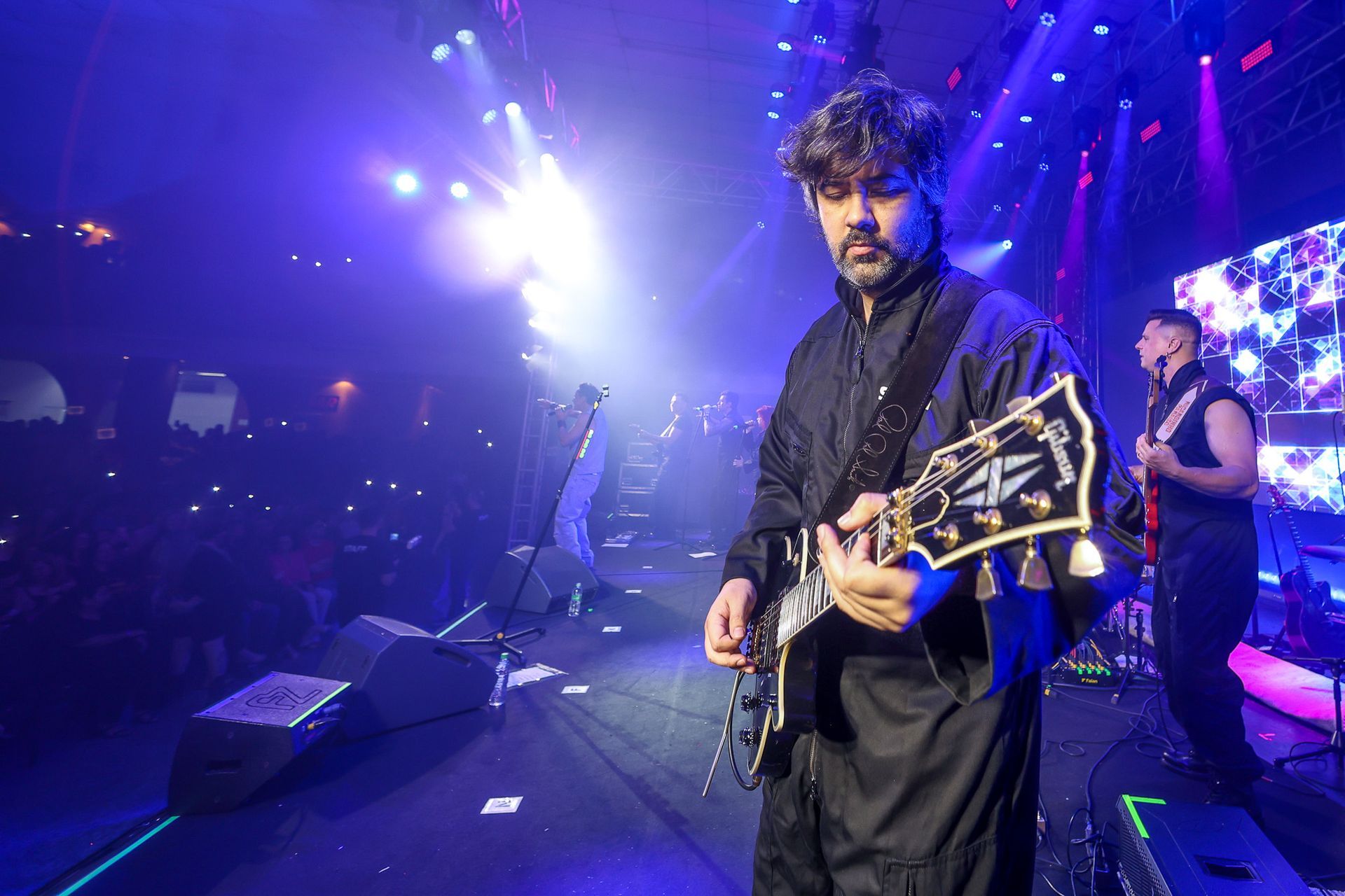 Um músico toca guitarra elétrica em um palco banhado por luzes azuis, com outro membro da banda visível nas proximidades.
