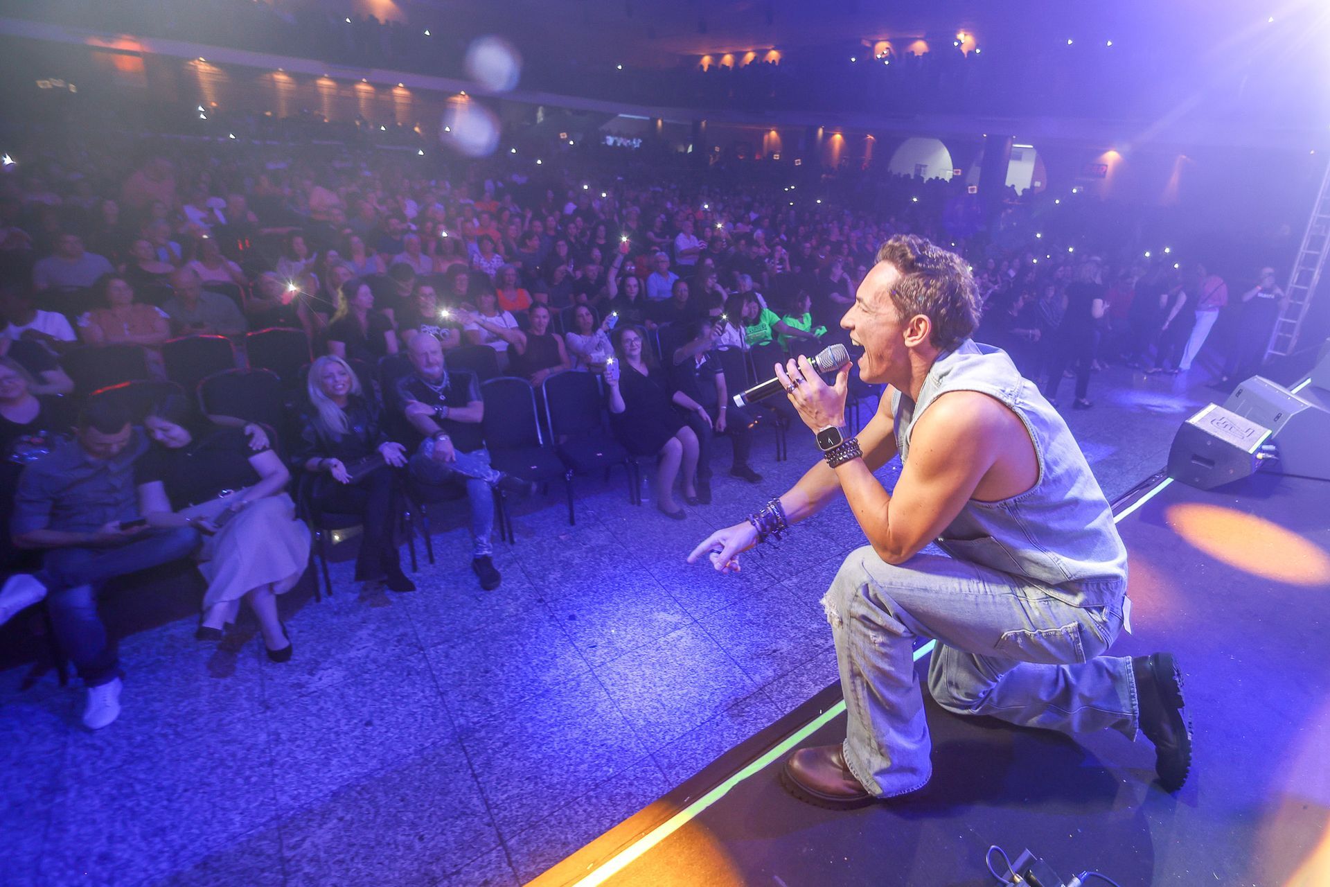 Um artista se ajoelha no palco, cantando diante de uma grande plateia com iluminação tênue, porém reduzida pela luz do palco.
