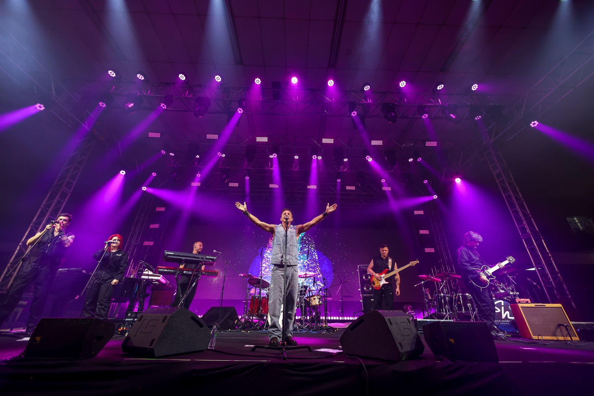 Uma banda se apresenta no palco sob luzes roxas, com o vocalista principal em pé no centro com os braços estendidos.