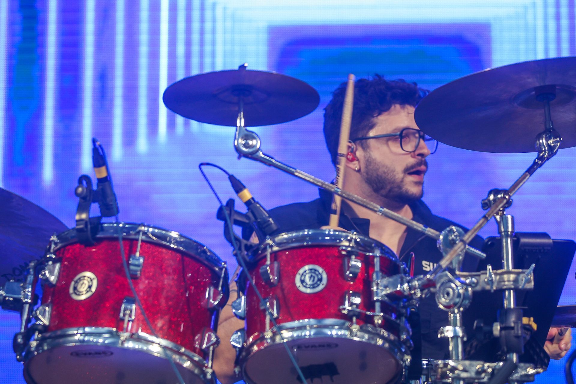 Um baterista de óculos se apresenta no palco, tocando uma bateria vermelha em frente a um painel de luzes azuis.
