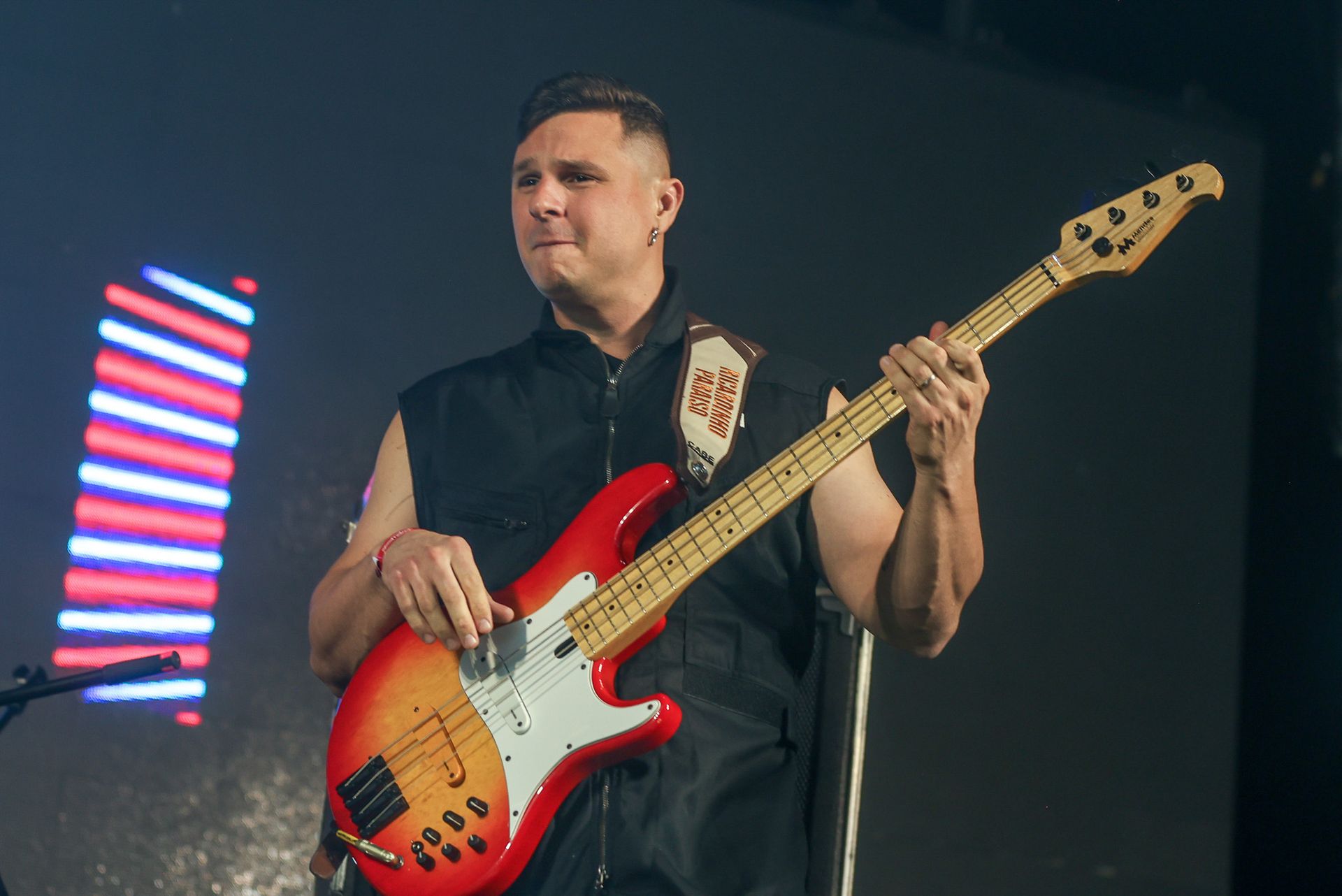 Um músico tocando um baixo vermelho e laranja em um palco pouco iluminado, com luzes vermelhas e azuis brilhantes atrás dele.