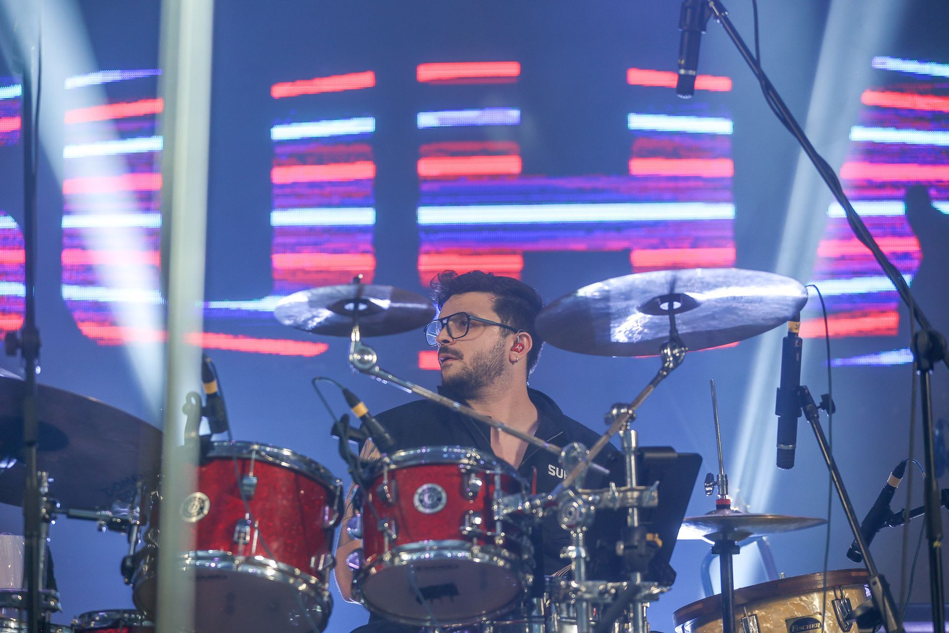 Um baterista de óculos tocando uma bateria vermelha no palco, em frente a um painel de LED