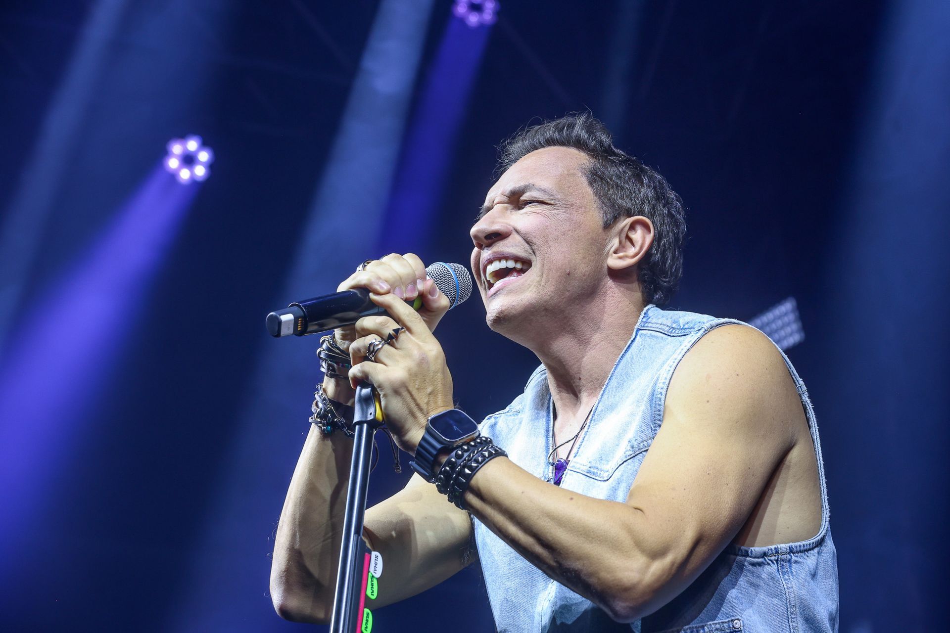 Uma pessoa vestindo uma camisa jeans sem mangas cantando com paixão em um microfone em um palco com iluminação azul de show.