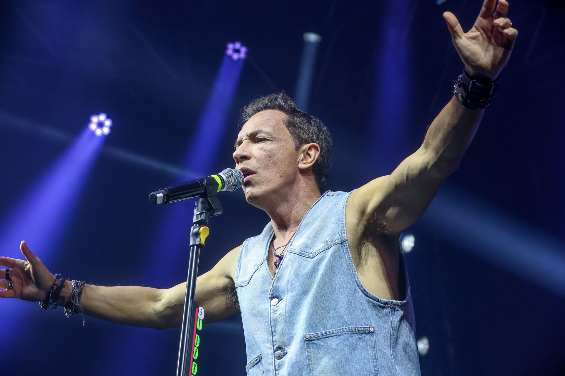 Um músico de colete jeans canta em um microfone no palco, com os braços estendidos sob holofotes azuis brilhantes.