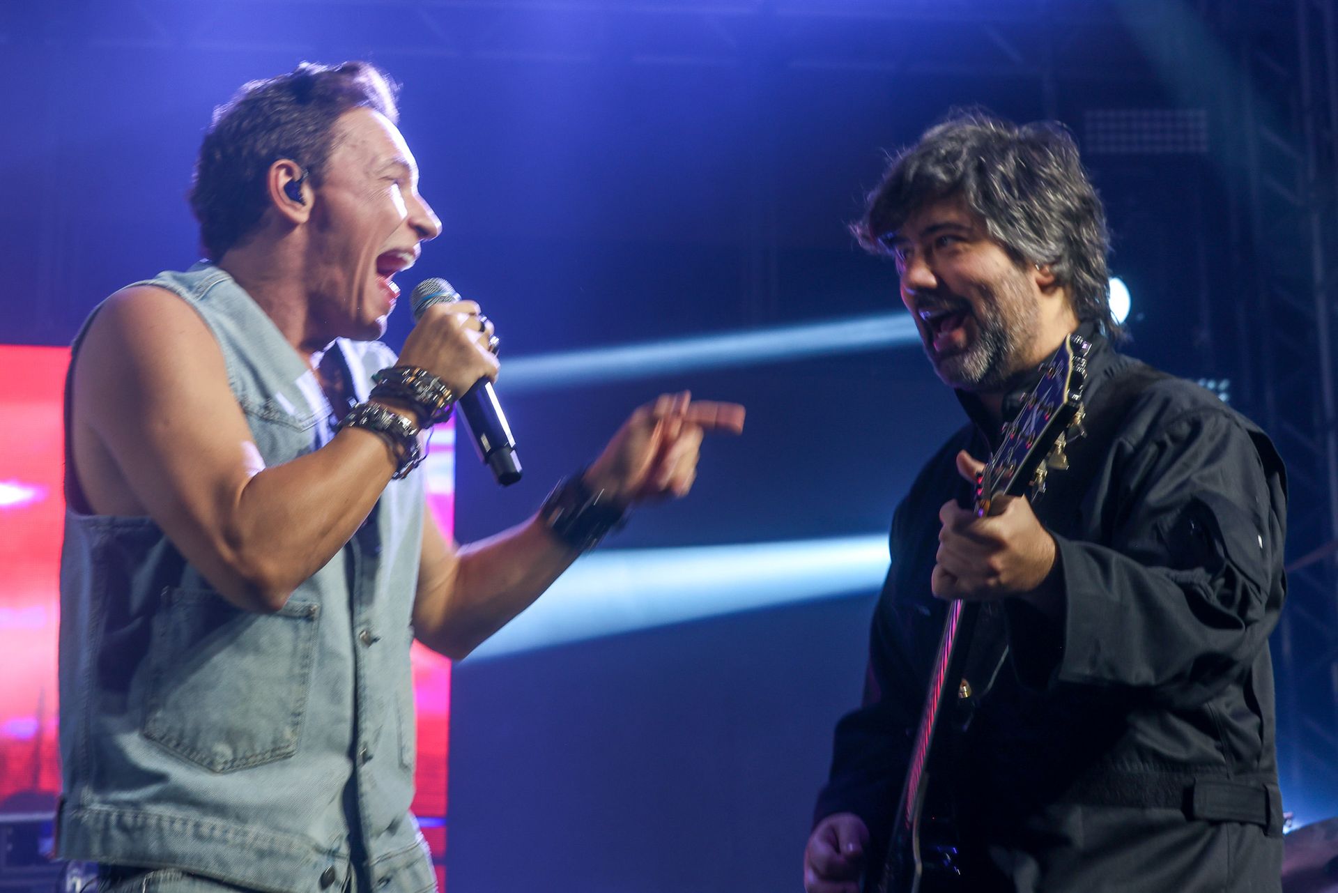 Um cantor de colete jeans e um guitarrista de jaqueta preta riem enquanto se apresentam juntos em um palco bem iluminado.