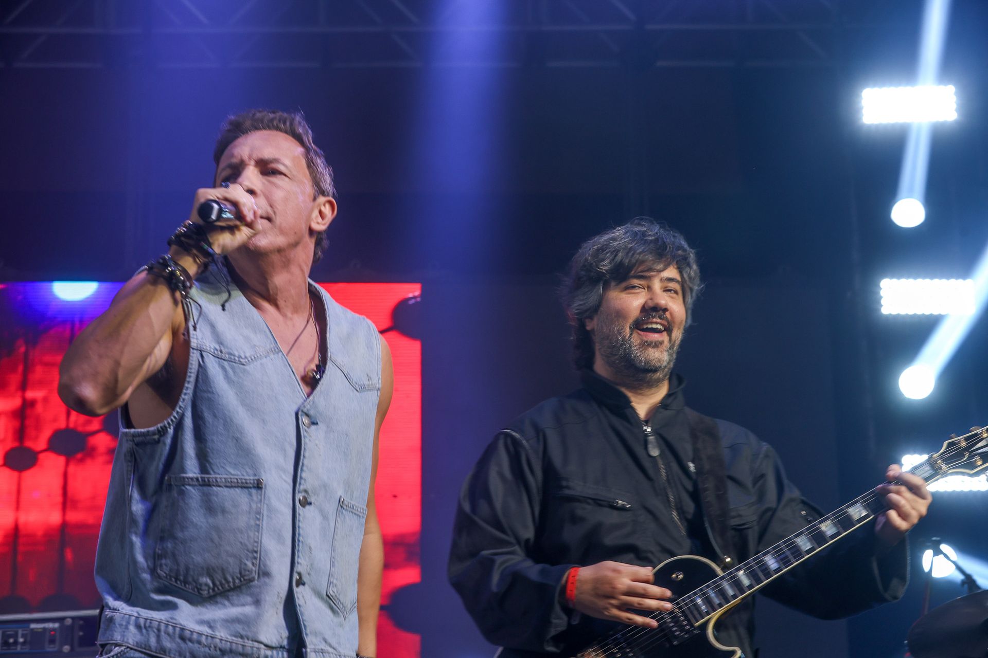 Dois músicos se apresentando no palco: um canta ao microfone enquanto o outro toca guitarra elétrica.