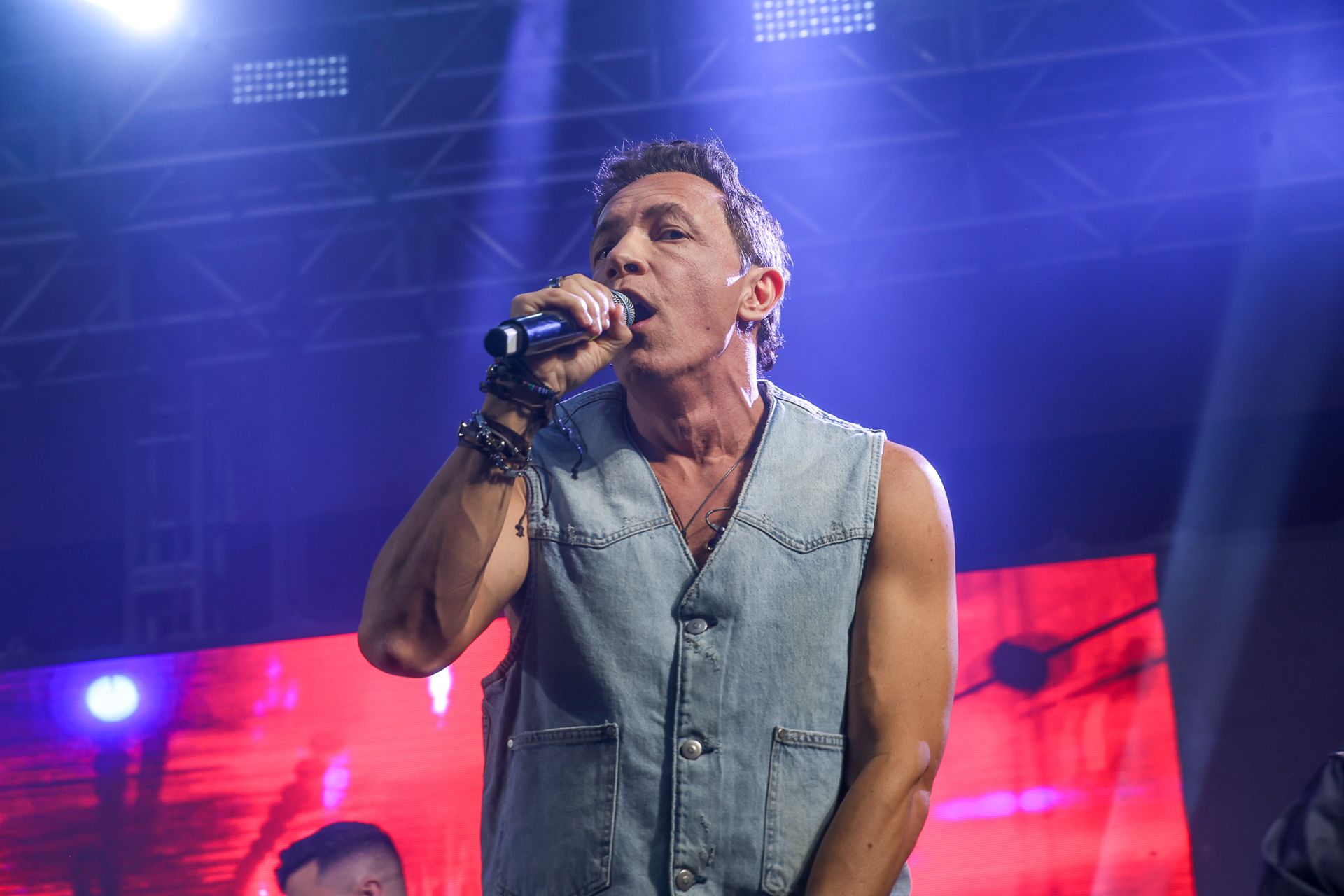Um artista vestindo um colete jeans canta em um microfone em um palco com iluminação brilhante em tons de roxo e vermelho.