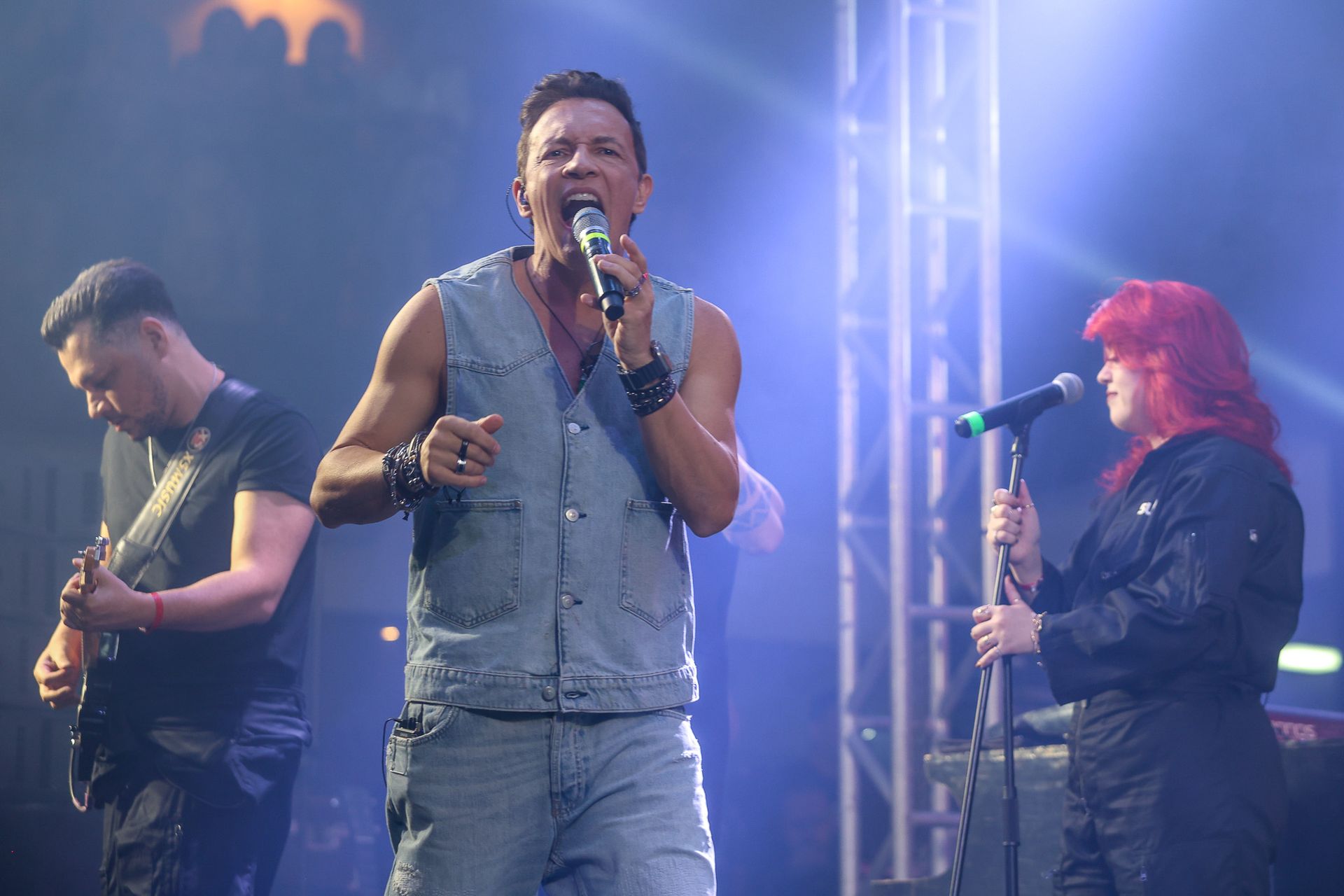 Uma banda se apresenta no palco sob luzes azuis: um cantor, um guitarrista e uma vocalista ruiva.