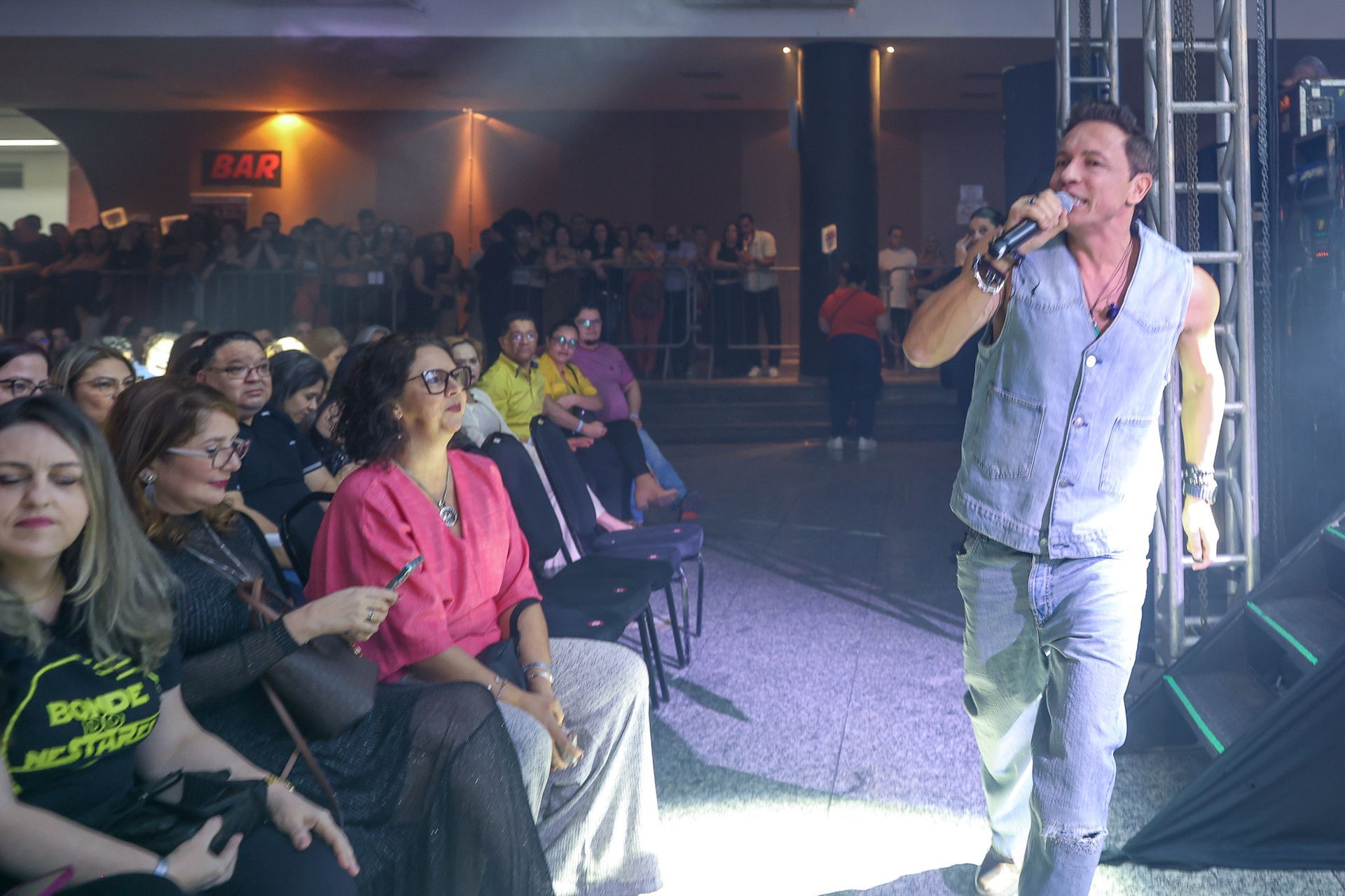 Um artista canta em um microfone no palco diante de uma plateia sentada em um local com pouca luz