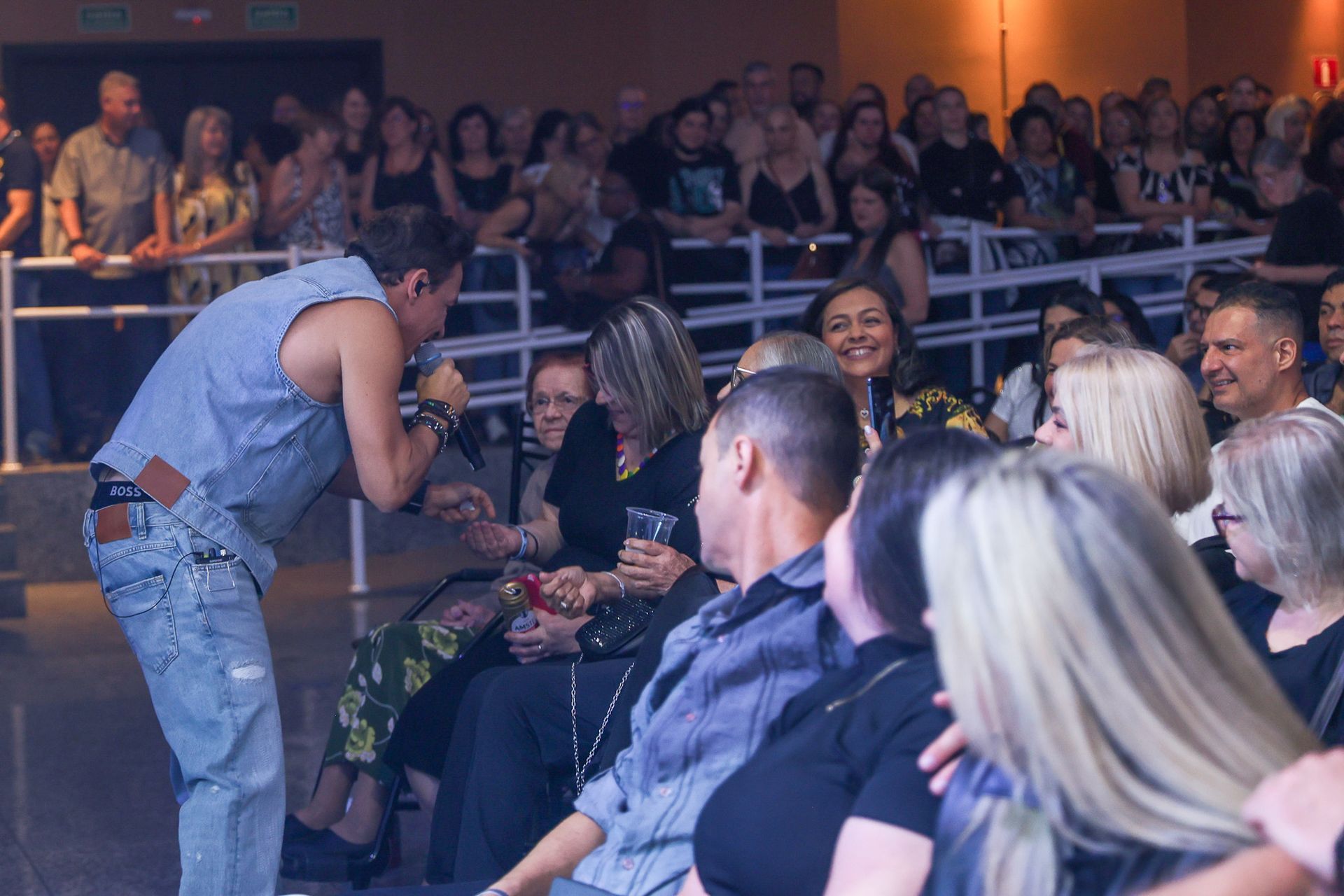 Um cantor vestindo colete e calça jeans se apresenta para uma plateia sentada em um grande local fechado.