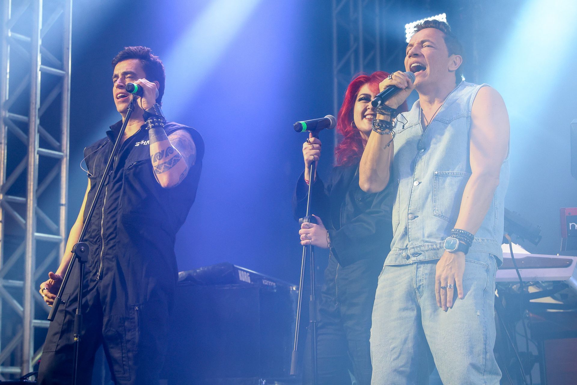 Três músicos se apresentam no palco sob holofotes azuis, cantando em microfones.