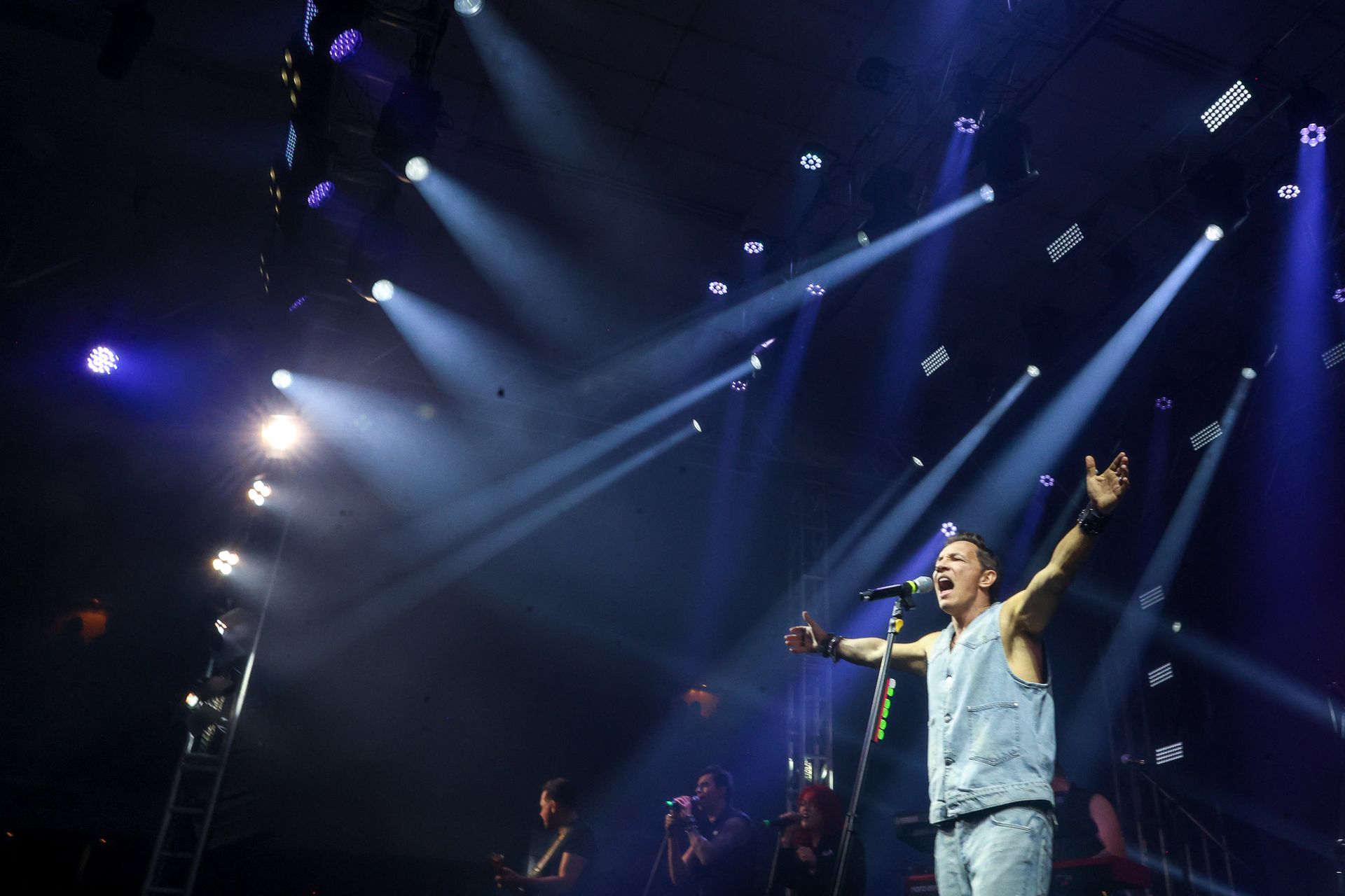 Um artista está em um palco com os braços estendidos, cantando em um microfone sob luzes de palco brilhantes.