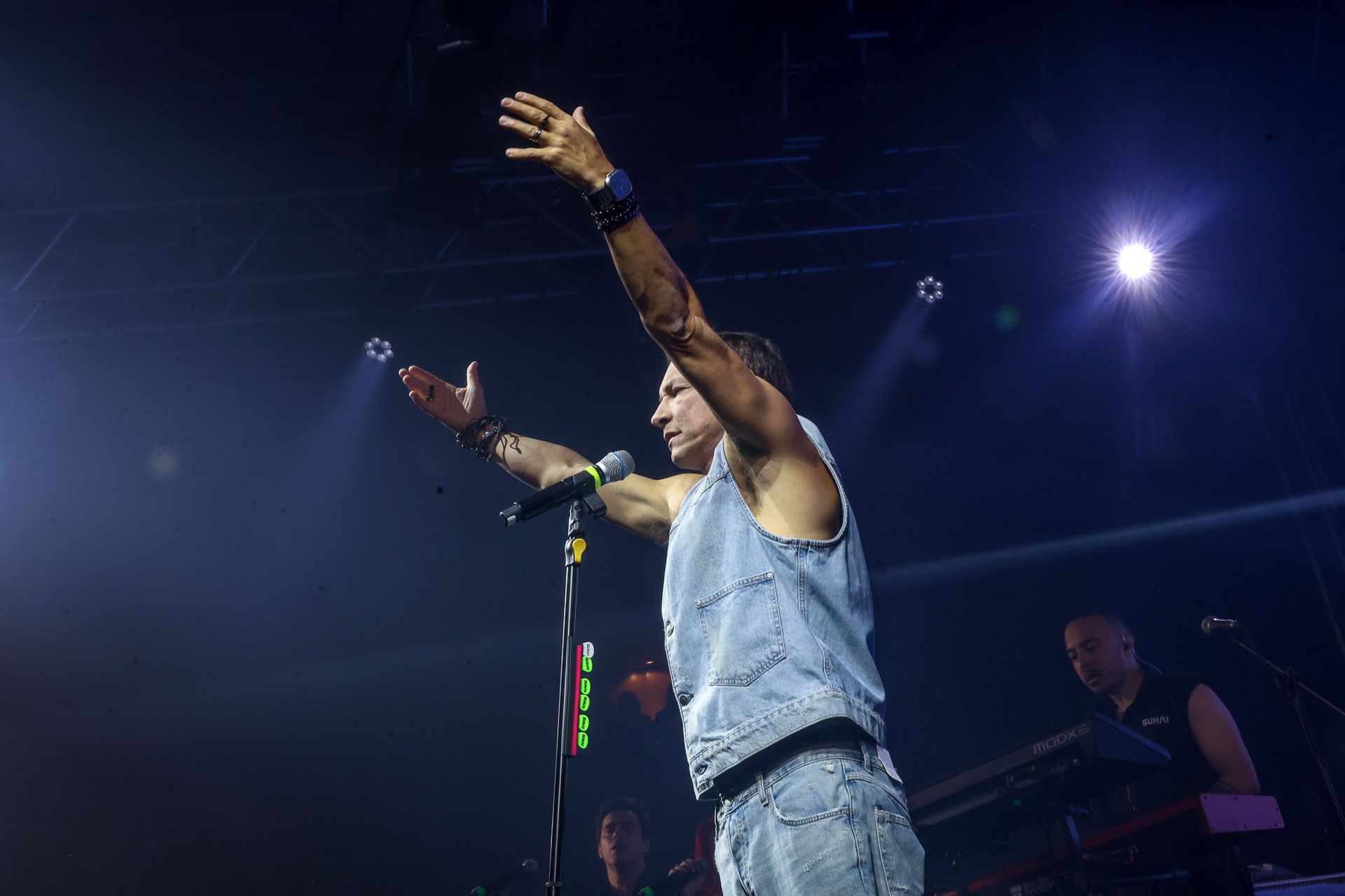 Um músico de colete jeans se apresenta no palco com os braços erguidos, segurando um microfone sob holofotes brilhantes.