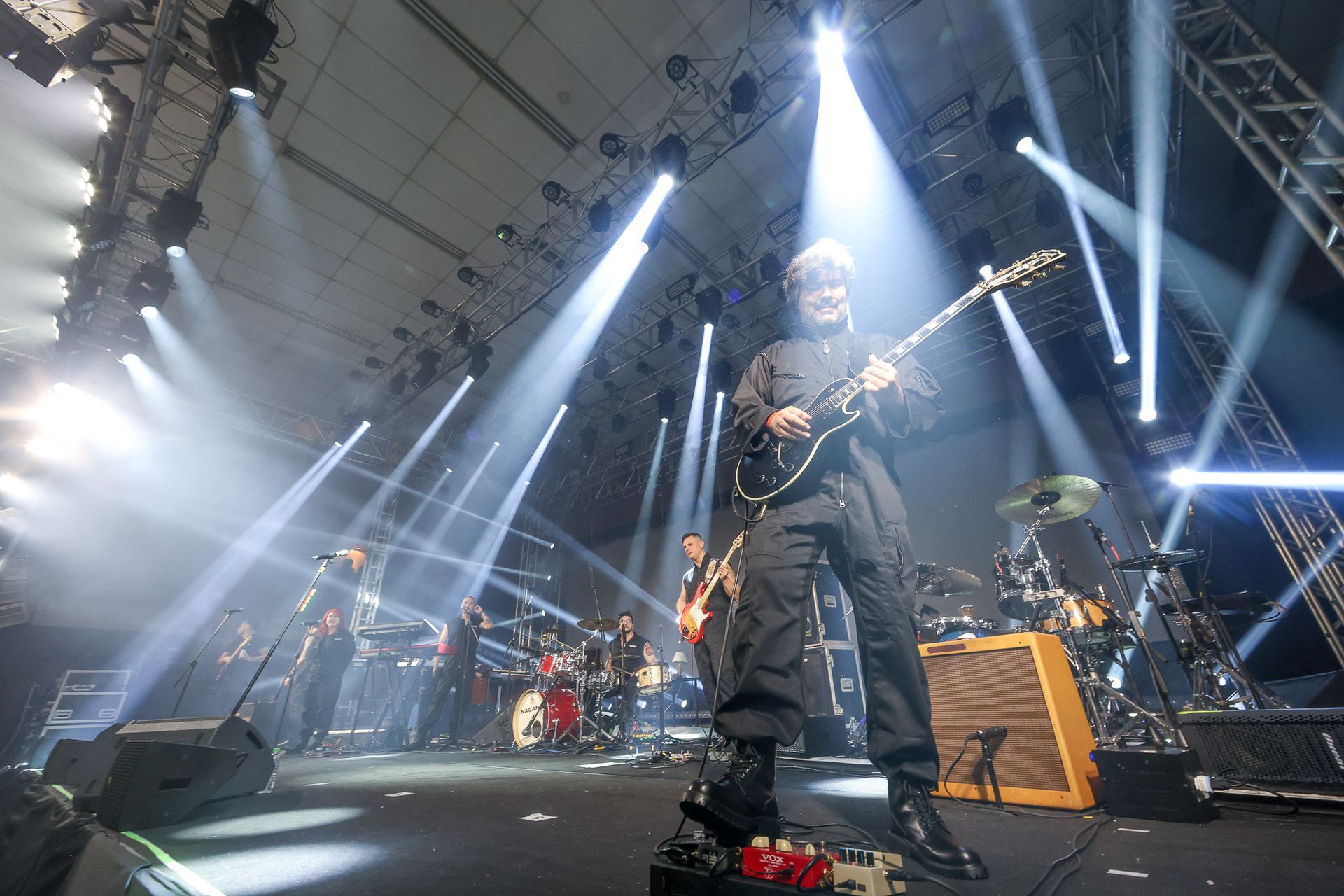 Uma banda de rock se apresenta em um palco de shows iluminado por holofotes brilhantes, com um guitarrista em primeiro plano.