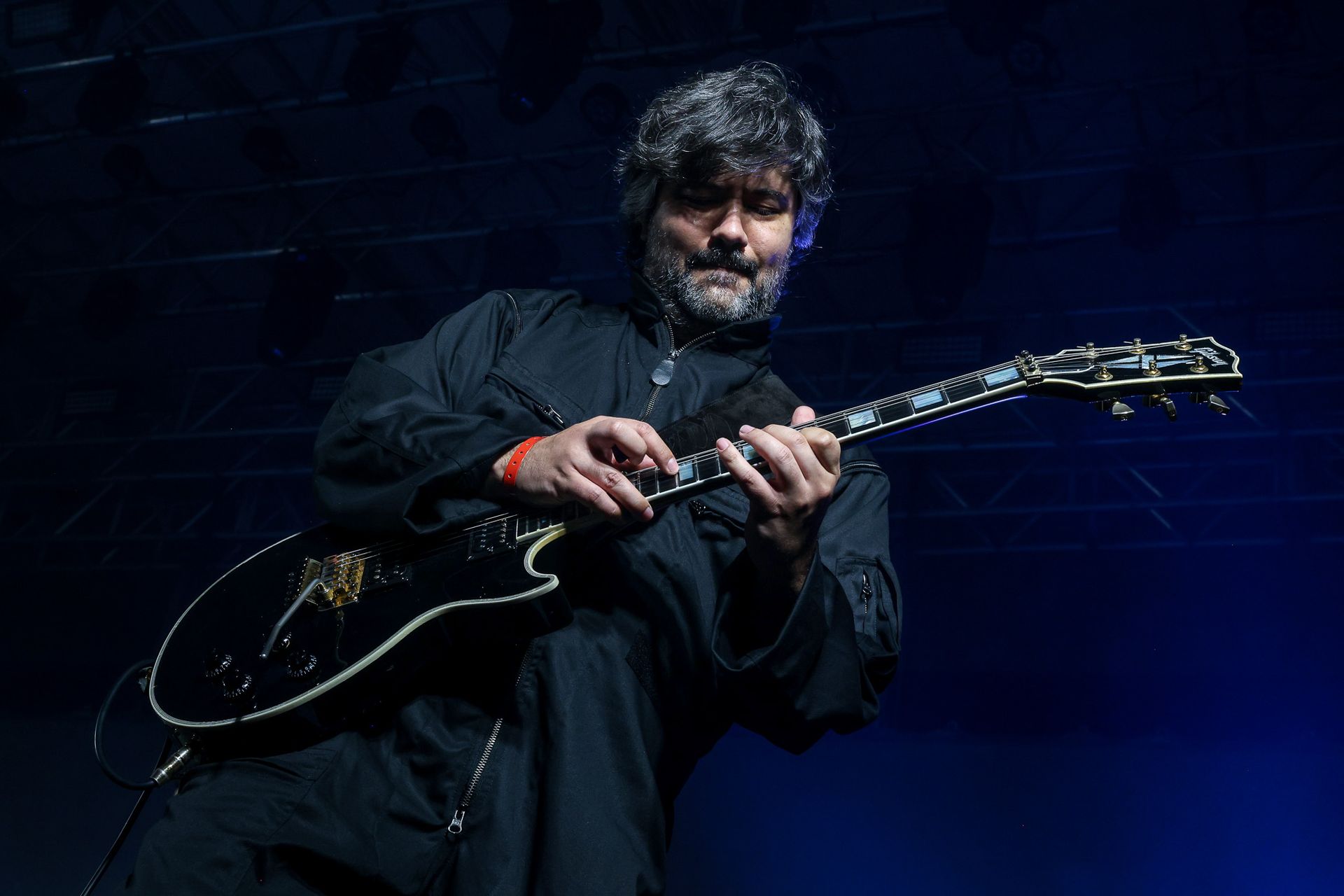 Um guitarrista barbudo tocando uma guitarra elétrica preta no palco sob iluminação azul escura.