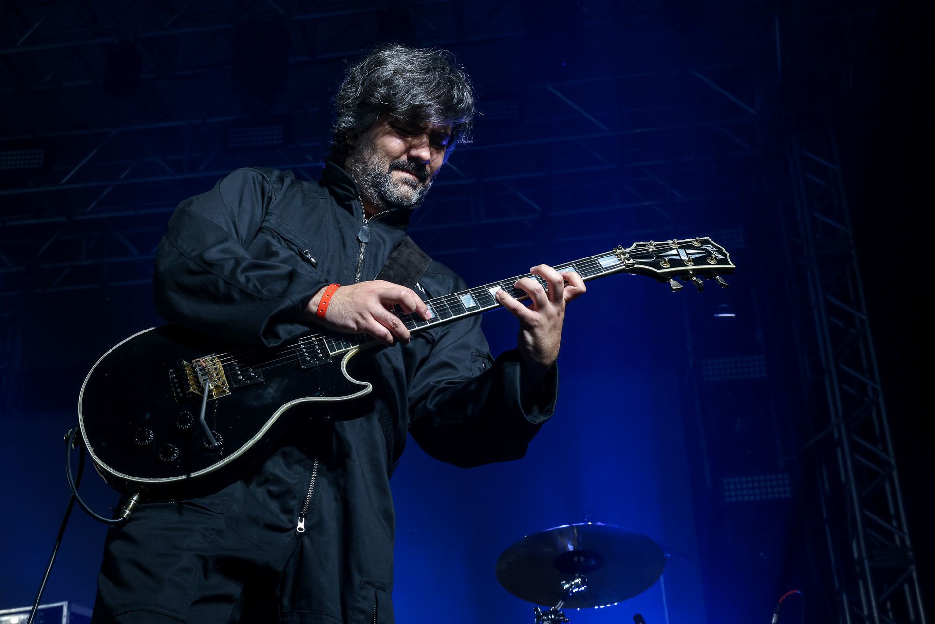 Um artista com cabelo despenteado e roupas escuras toca uma guitarra elétrica preta em um palco pouco iluminado com luz azul.