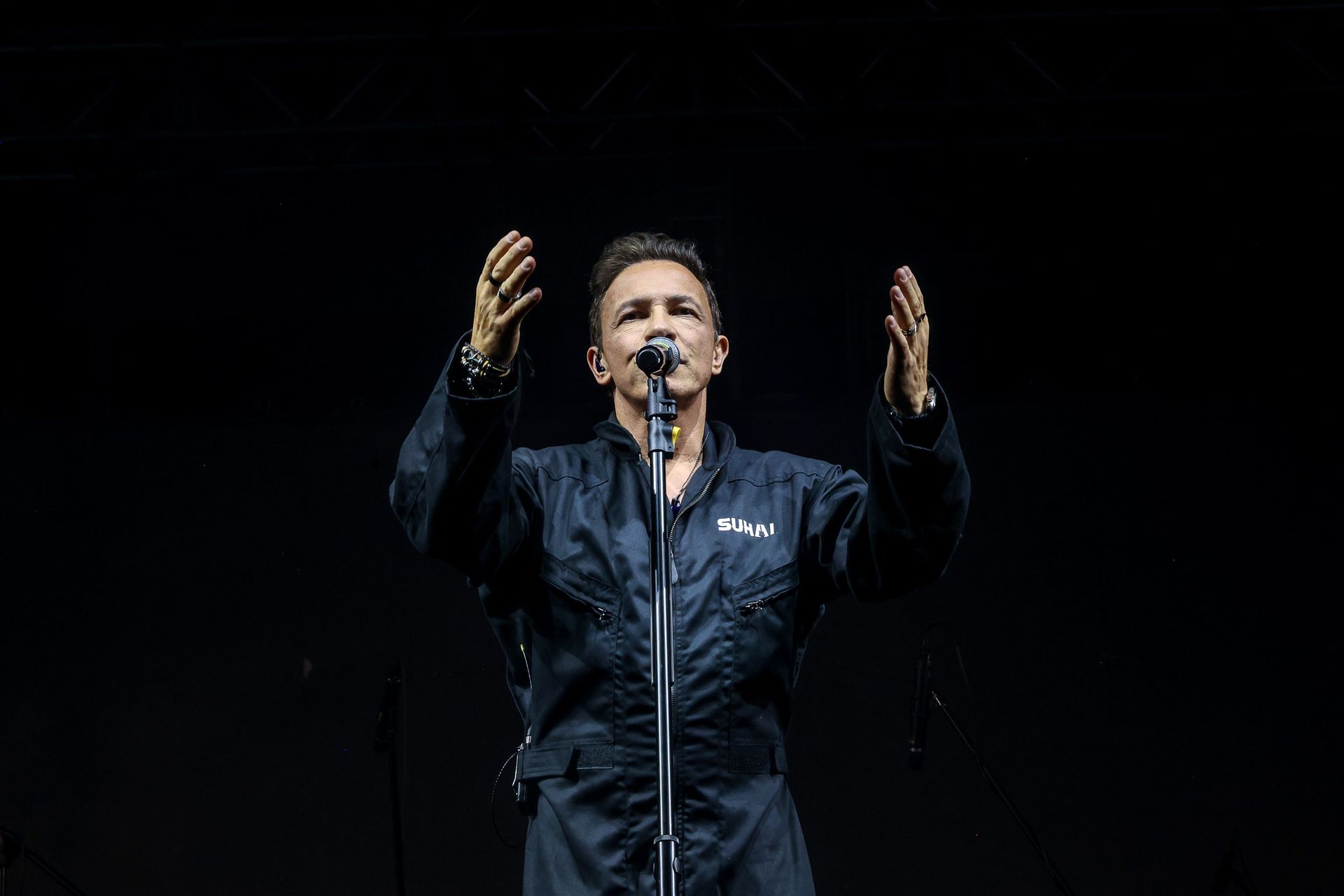 Uma artista de macacão preto está no centro do palco sob um holofote, com as mãos erguidas, cantando em um microfone.