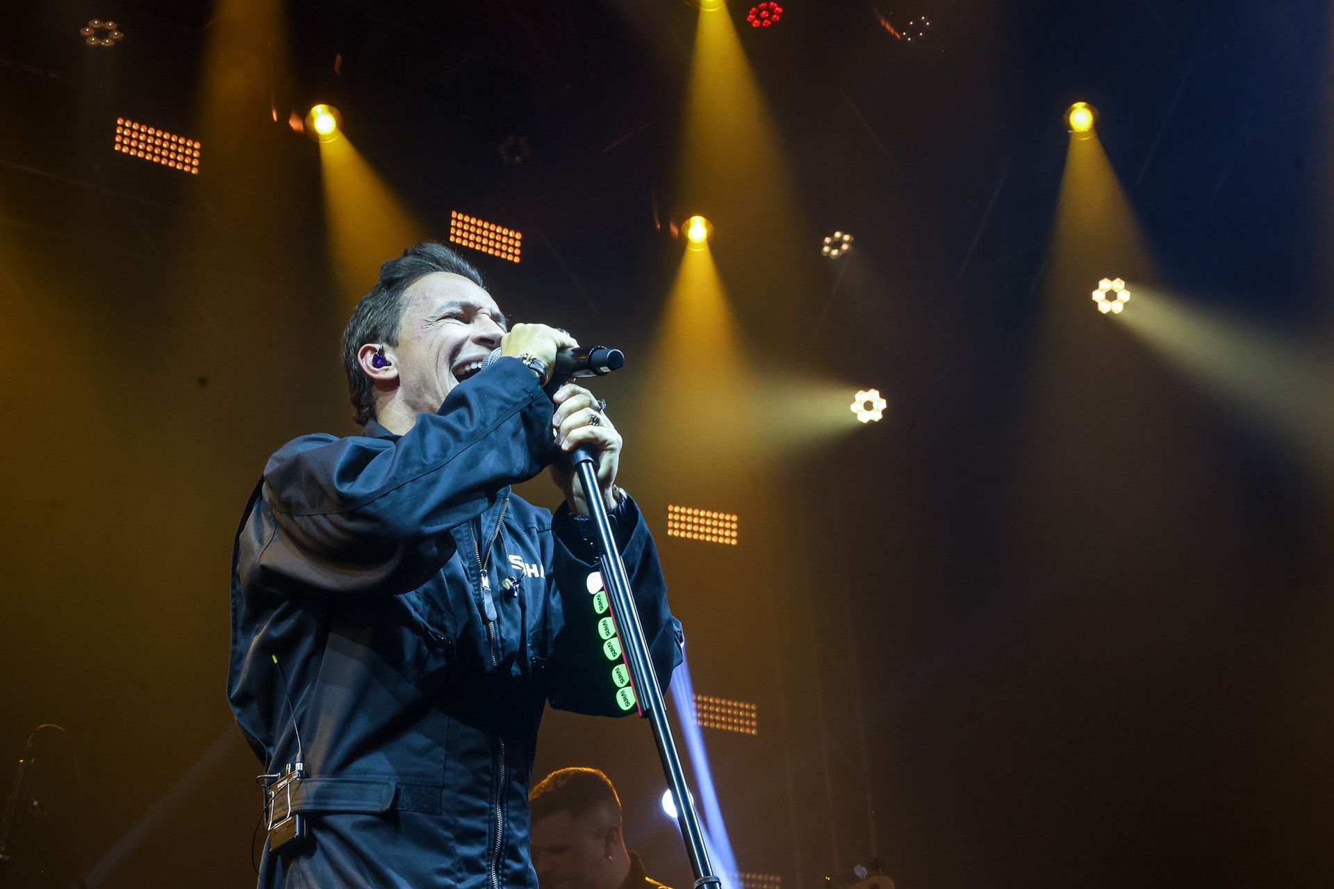 Um artista canta em um microfone em um palco iluminado por vários holofotes brilhantes contra um fundo escuro.