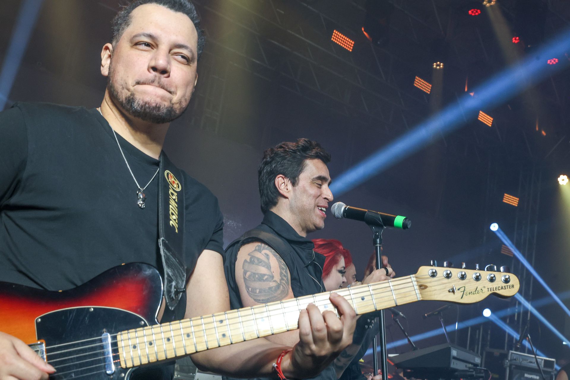 Dois músicos se apresentam no palco sob holofotes; um toca uma guitarra enquanto o outro canta ao microfone.