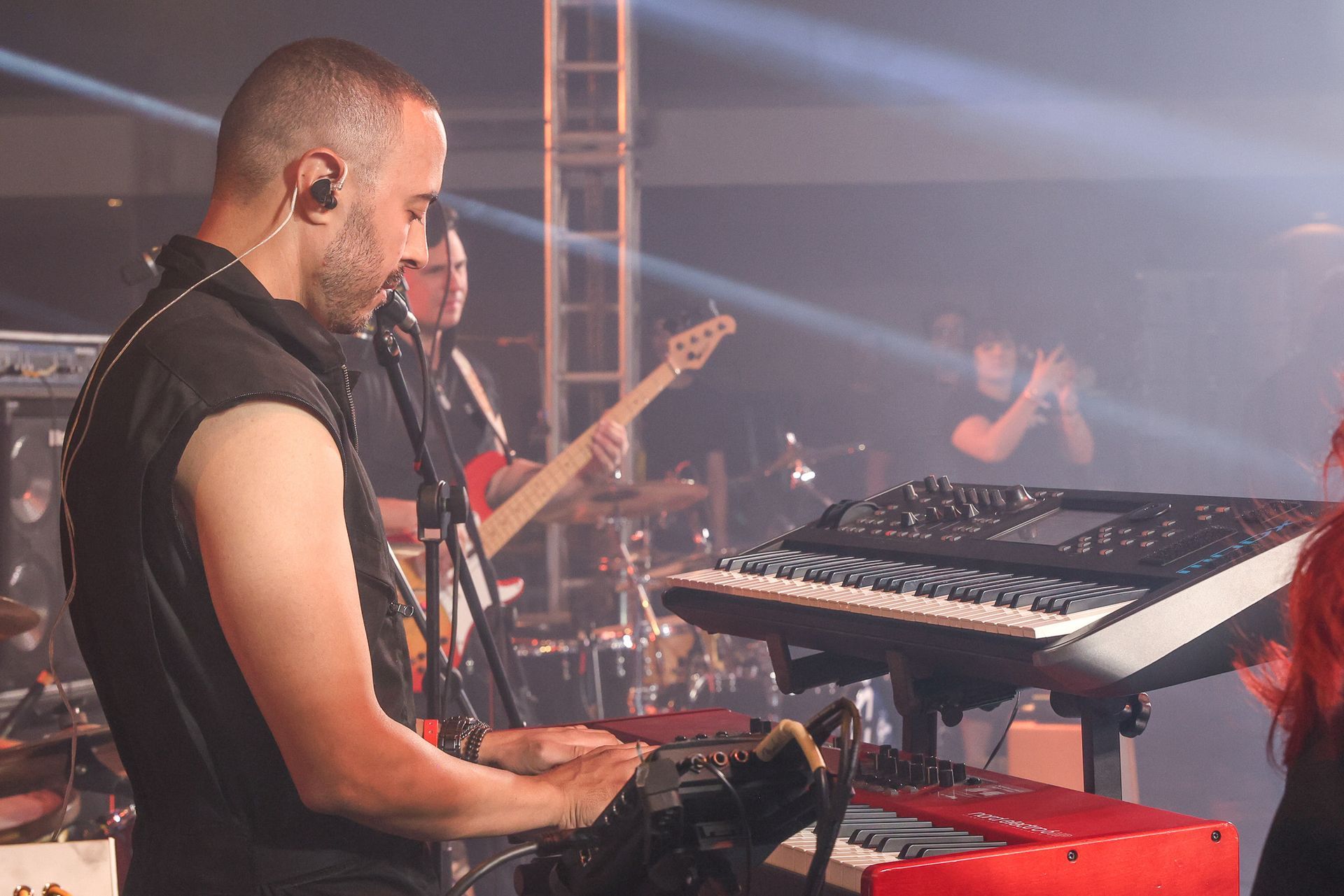 Um músico toca teclado no palco durante um concerto enquanto um baixista toca ao fundo sob a iluminação do palco.