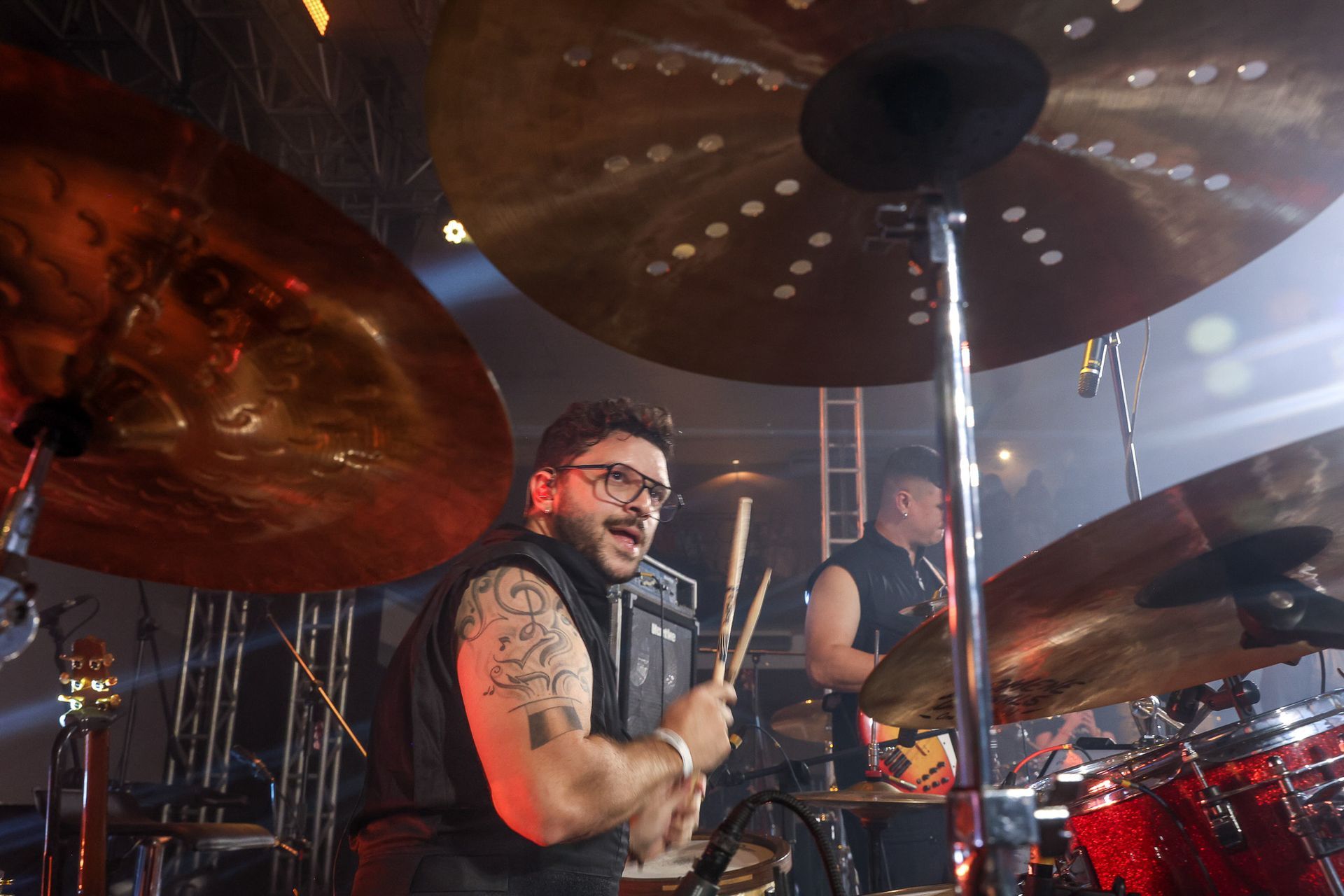 Um baterista de óculos e tatuagens toca bateria em um palco, com um segundo músico visível ao fundo desfocado.