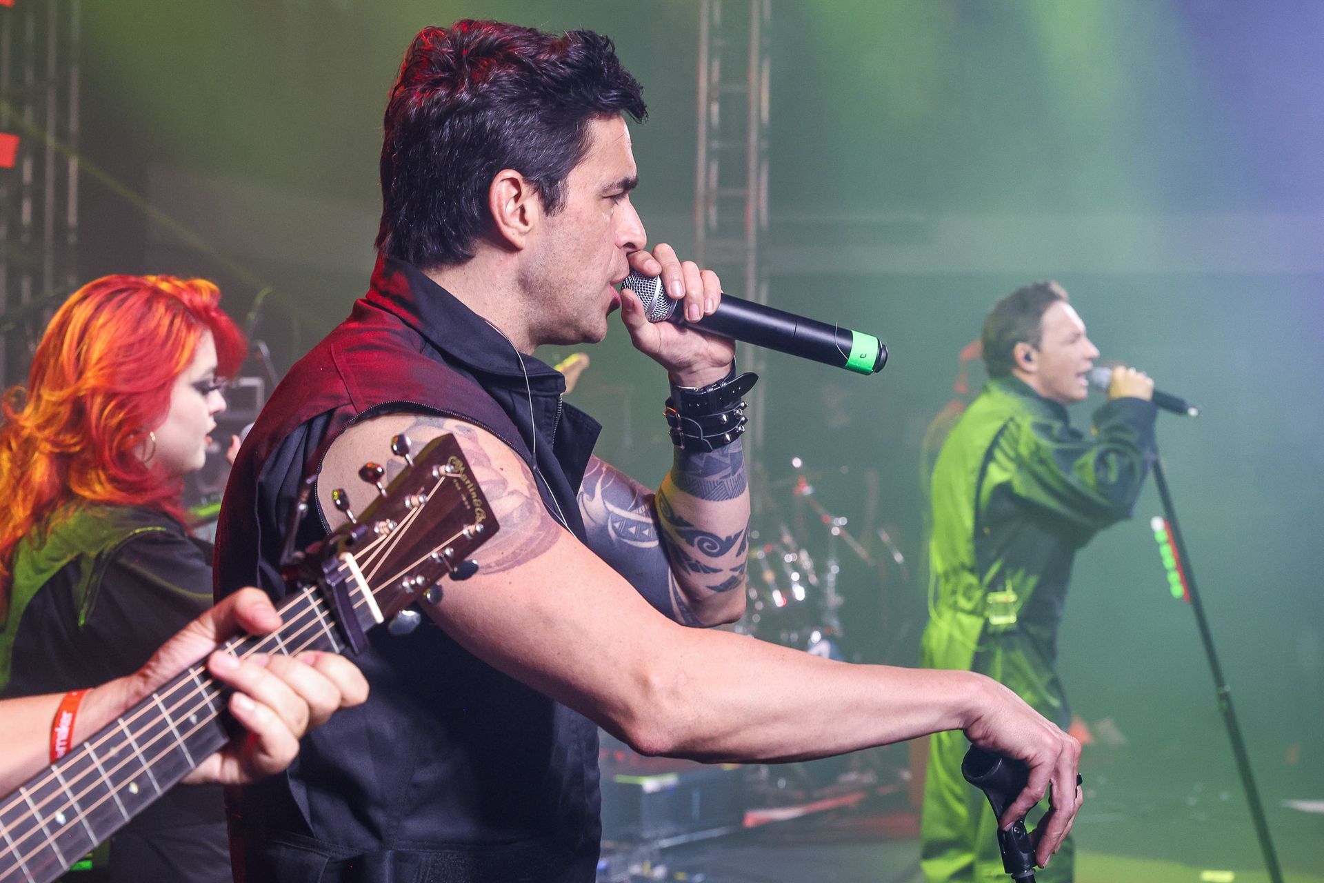 Uma banda se apresentando no palco, com o vocalista usando um microfone e um guitarrista tocando em primeiro plano.